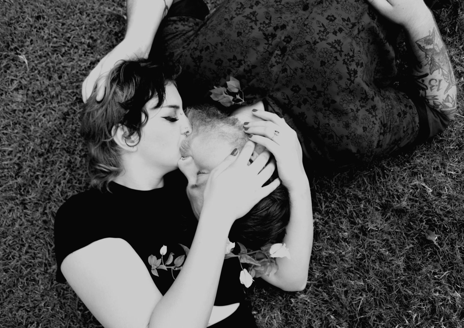 Black and white photo of two women lying on grass sharing a gentle kiss while embracing.