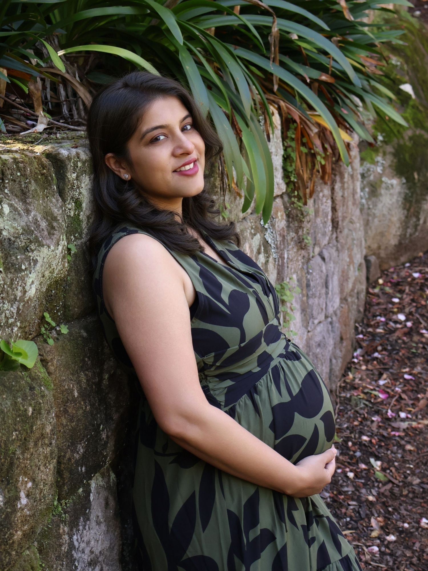 Smiling pregnant woman in green floral dress leaning against a stone wall in a garden.