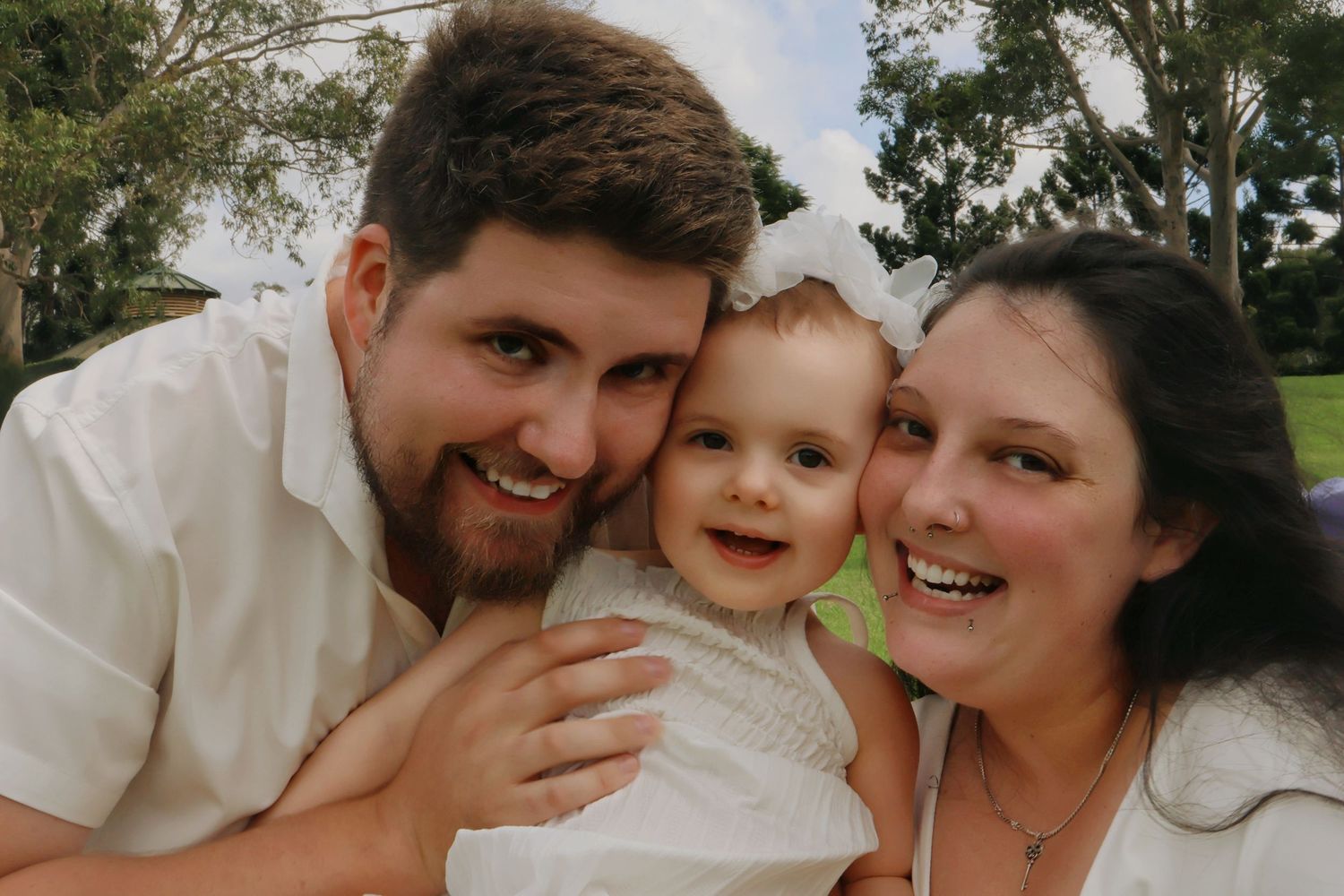 Happy young family with a smiling baby between two parents in white shirts posing outdoors in a park.