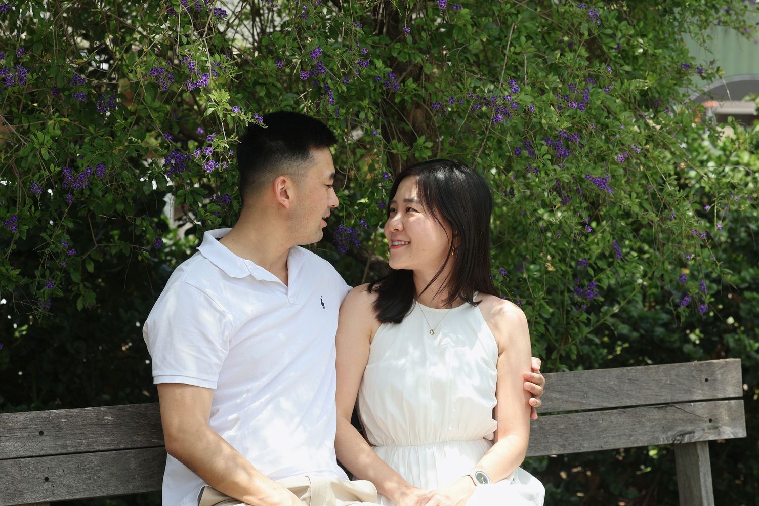 A couple sits on a bench in a garden with purple flowers, smiling warmly at each other.