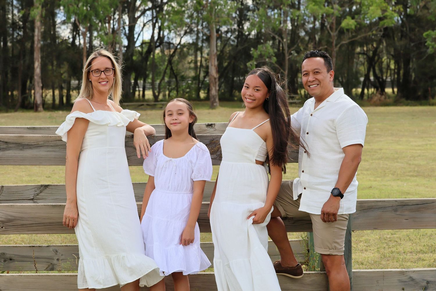 Joyful family of four posing in matching white outfits by a rustic wooden fence outdoors.