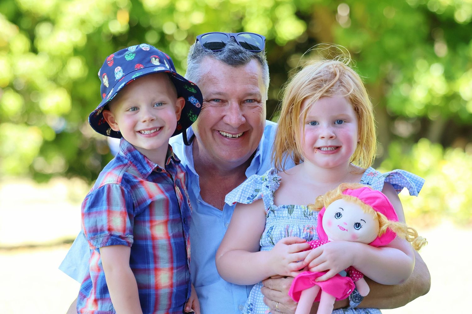 Smiling father poses outdoors with two young children and a colorful toy doll.