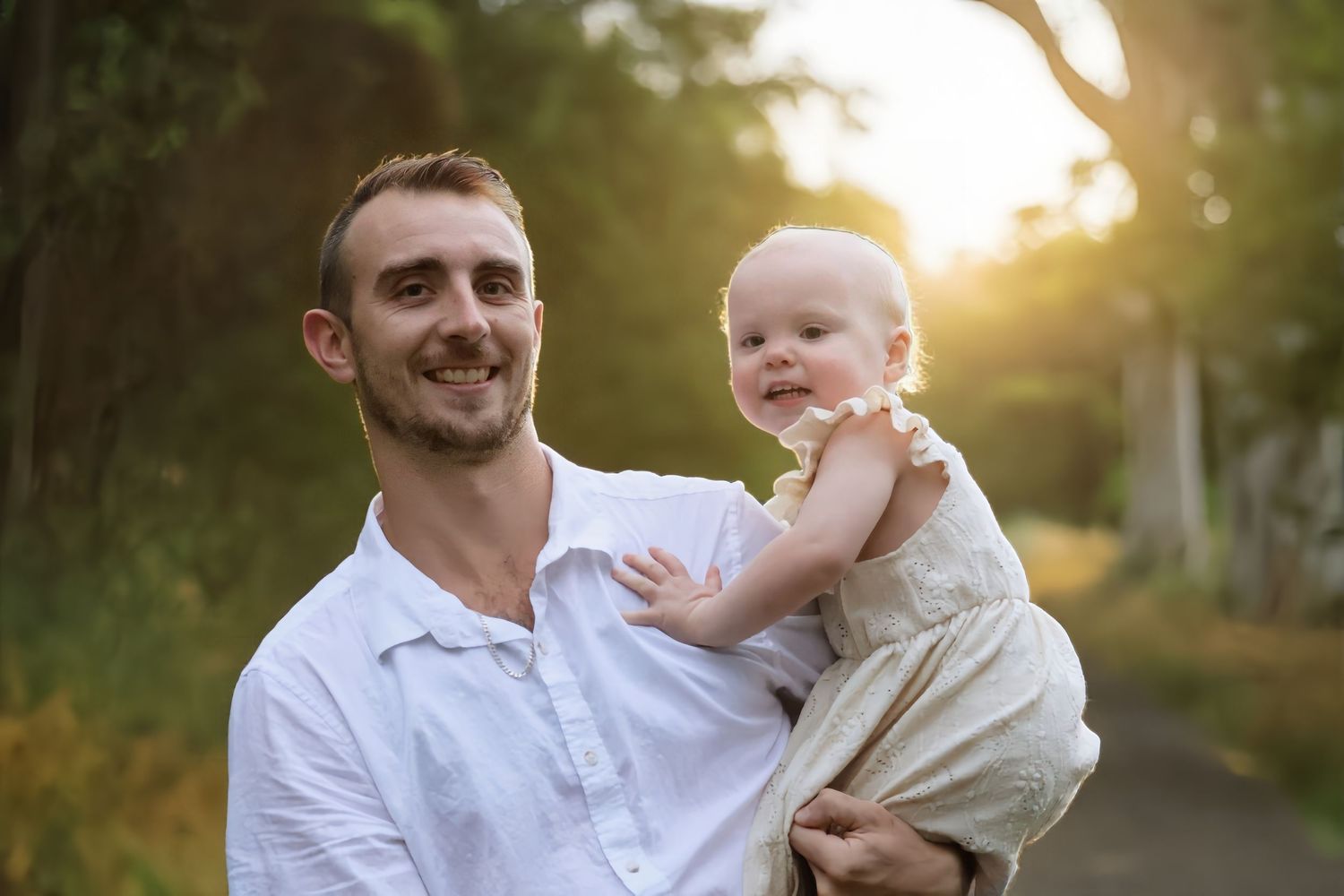 Smiling young father holding a happy baby girl outdoors in golden sunset light.