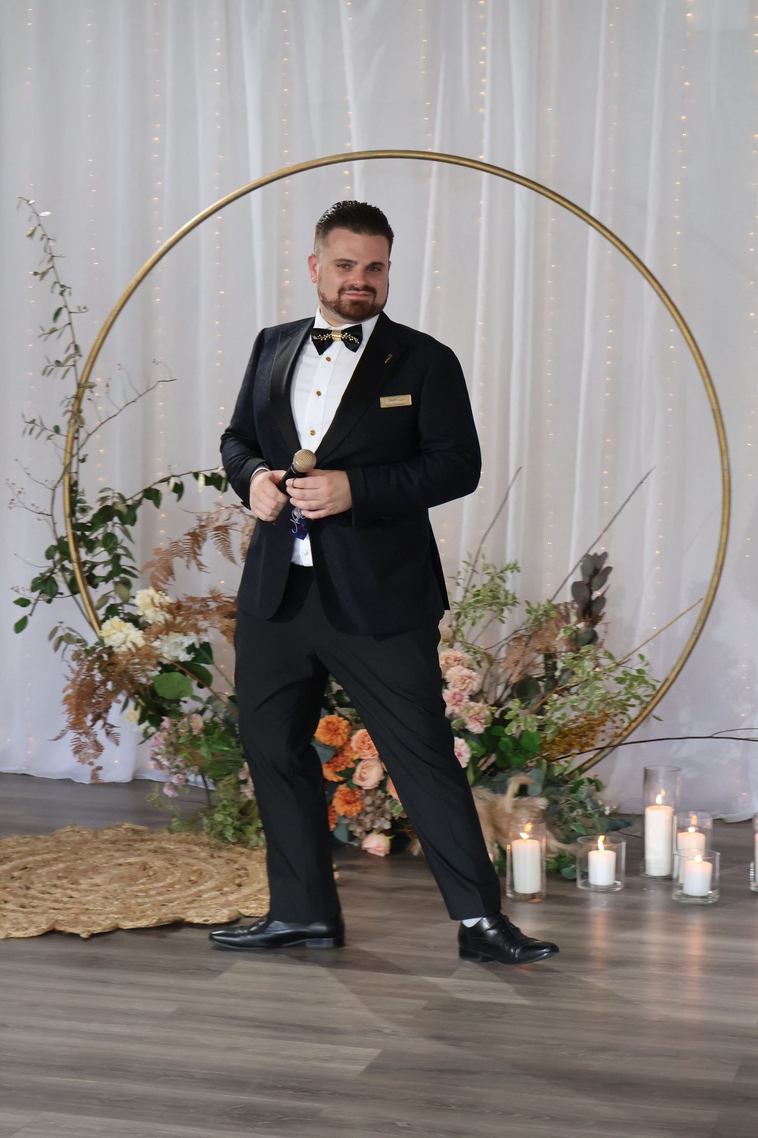 Groom in black tuxedo posing confidently in front of circular floral arch at wedding venue.