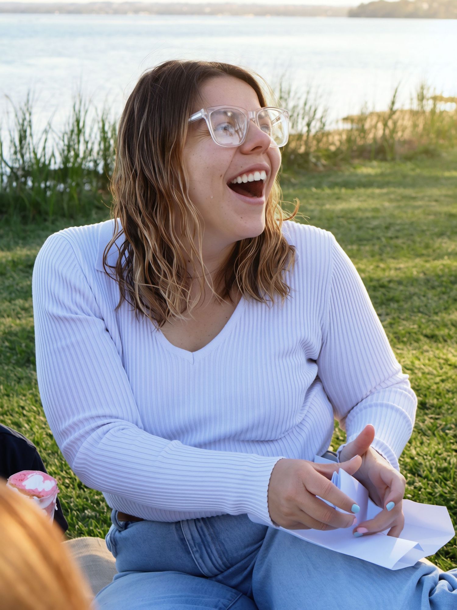 Woman with glasses laughing joyfully while sitting on grass near a waterfront on a sunny day.