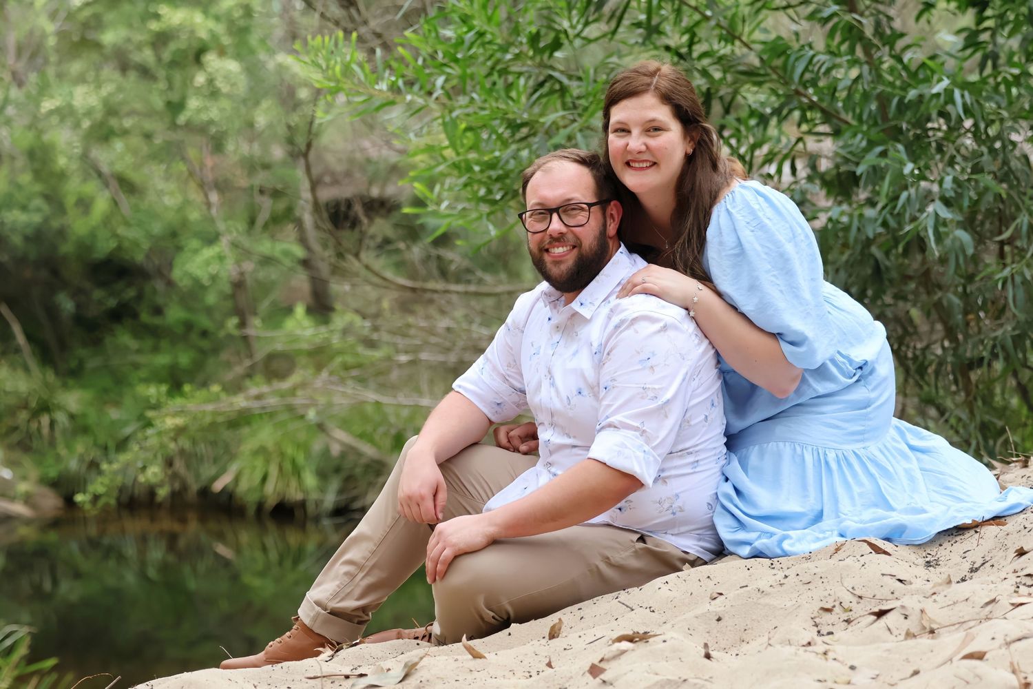 Smiling couple sits together on sandy riverbank surrounded by lush green trees and calm water.