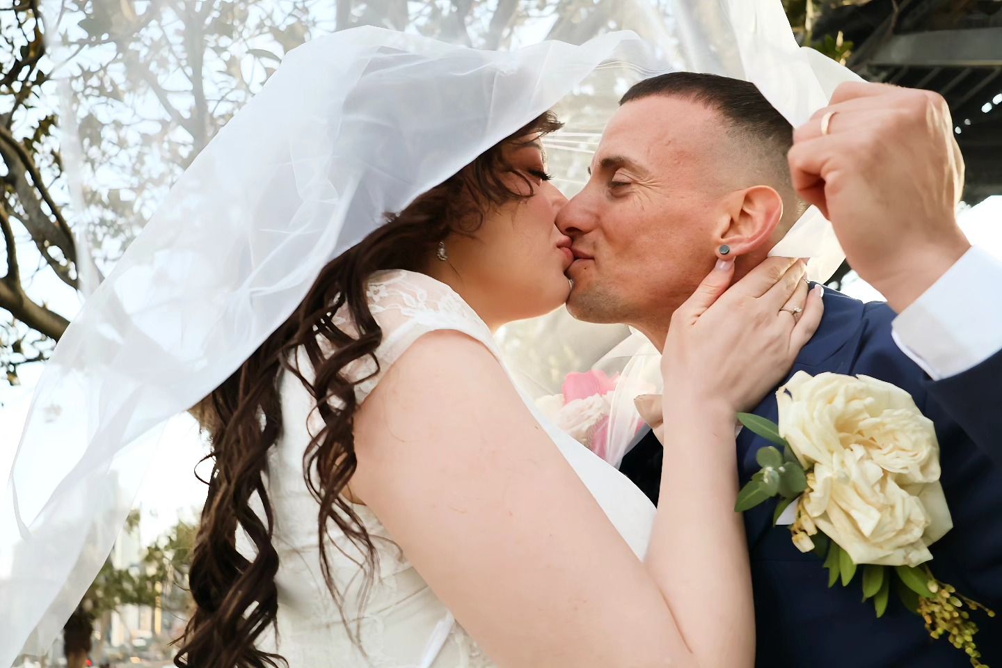Bride and groom share a kiss under a white veil, bride holding a white bouquet on their wedding day.