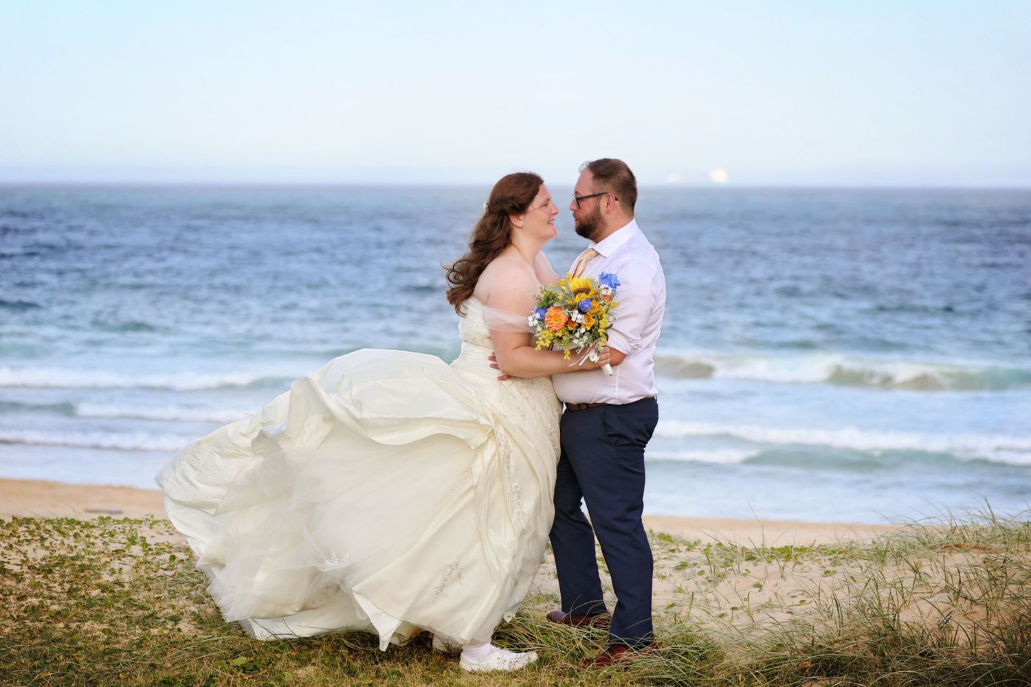 Bride and groom embracing on beach, wedding dress flowing in wind, holding colorful bouquet by ocean.
