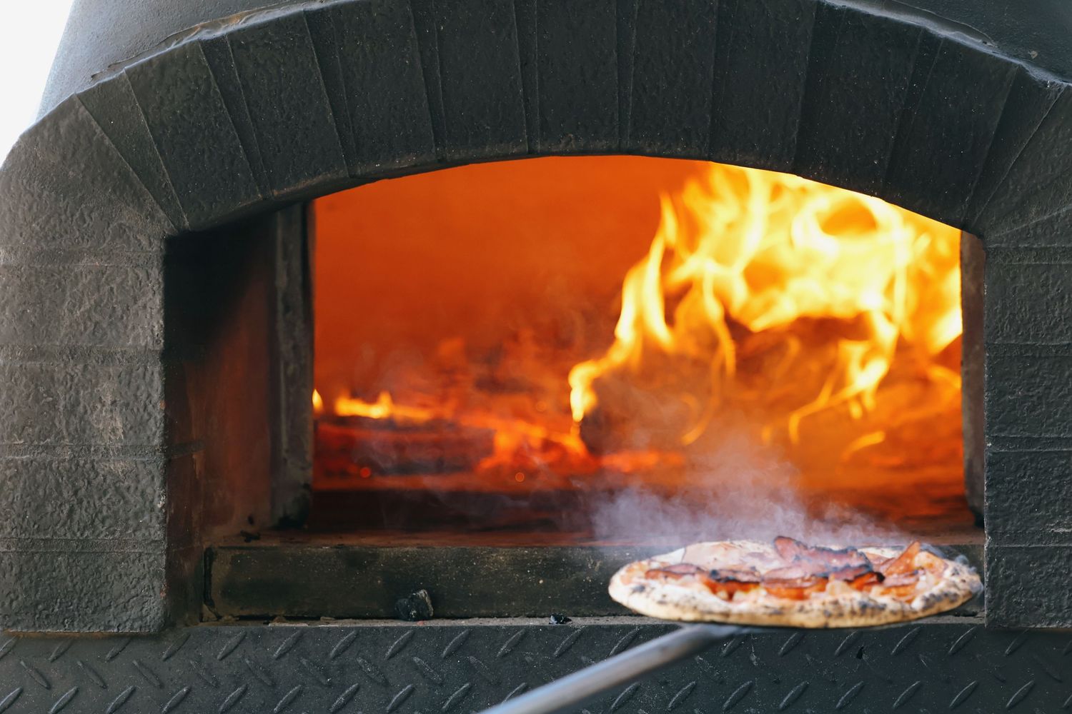 Pizza cooking in a wood-fired oven with open flames visible inside a stone arch.