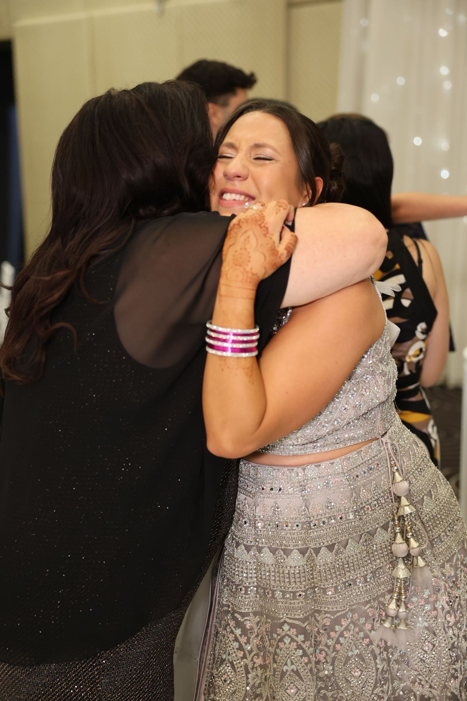 Emotional woman in silver lehenga receiving a heartfelt hug at a wedding celebration.