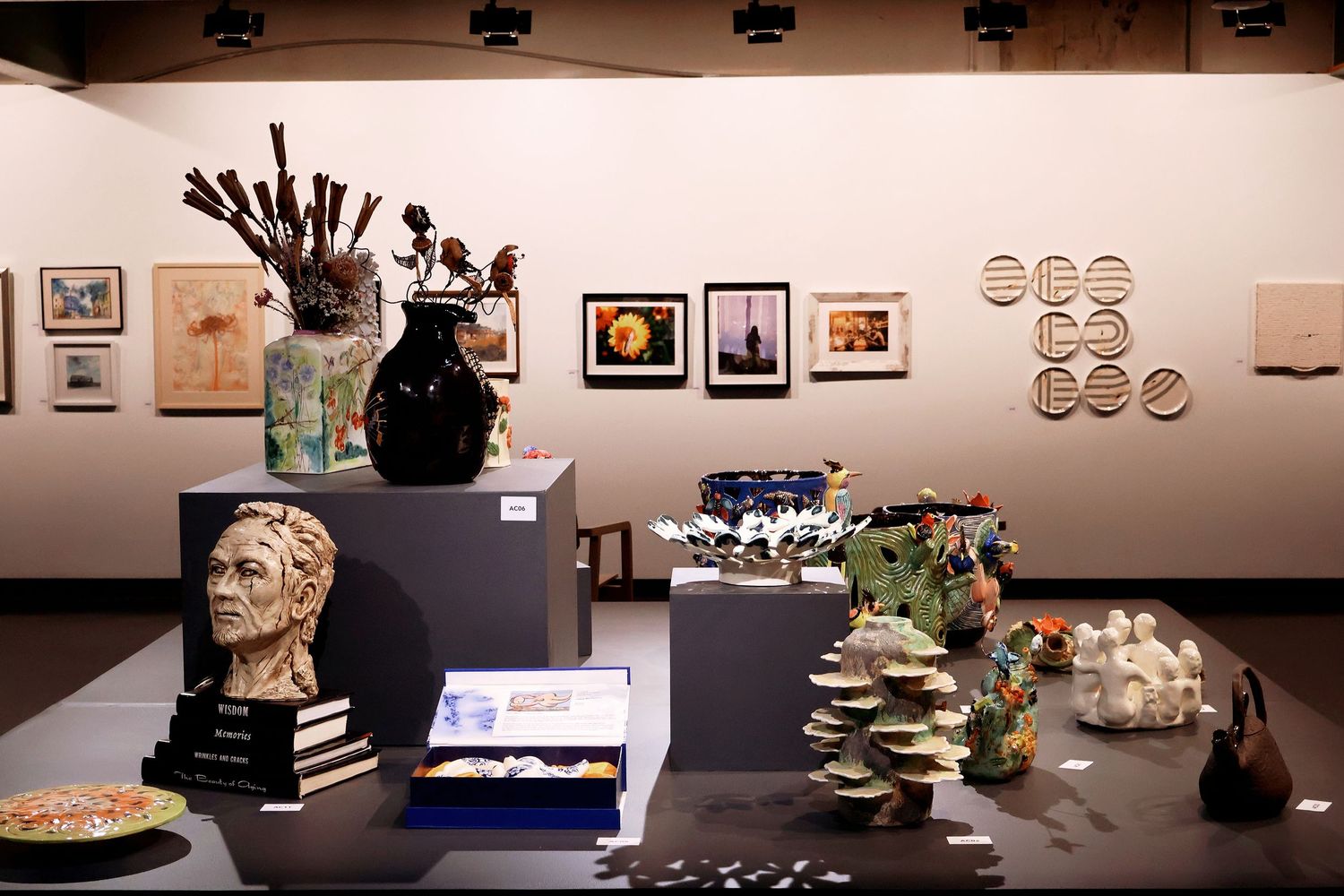Diverse art exhibition display featuring ceramic sculptures, bust, vase, and mixed media artworks in gallery.