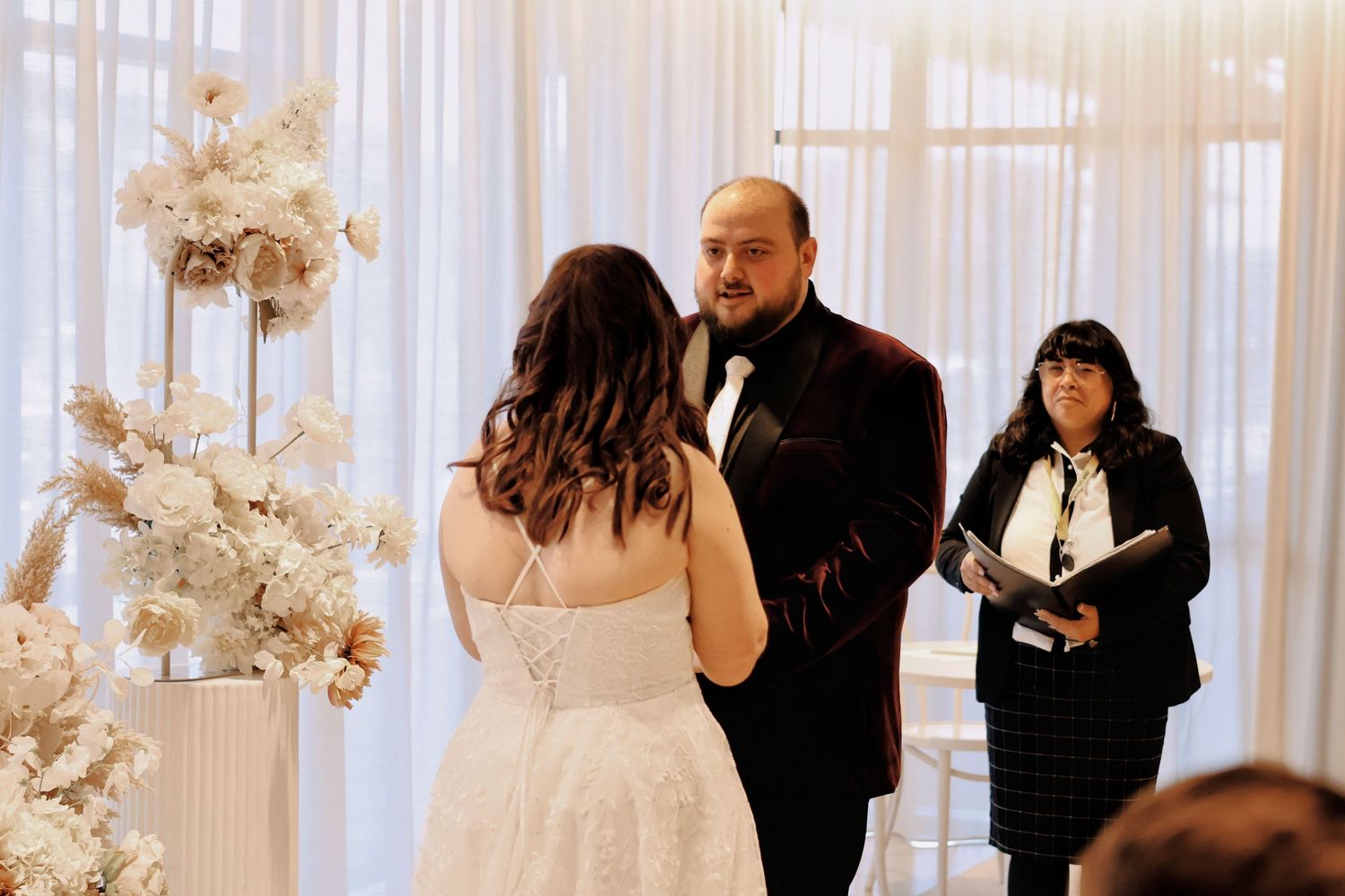 Groom reciting vows to bride at elegant white wedding ceremony with officiant and floral backdrop visible.