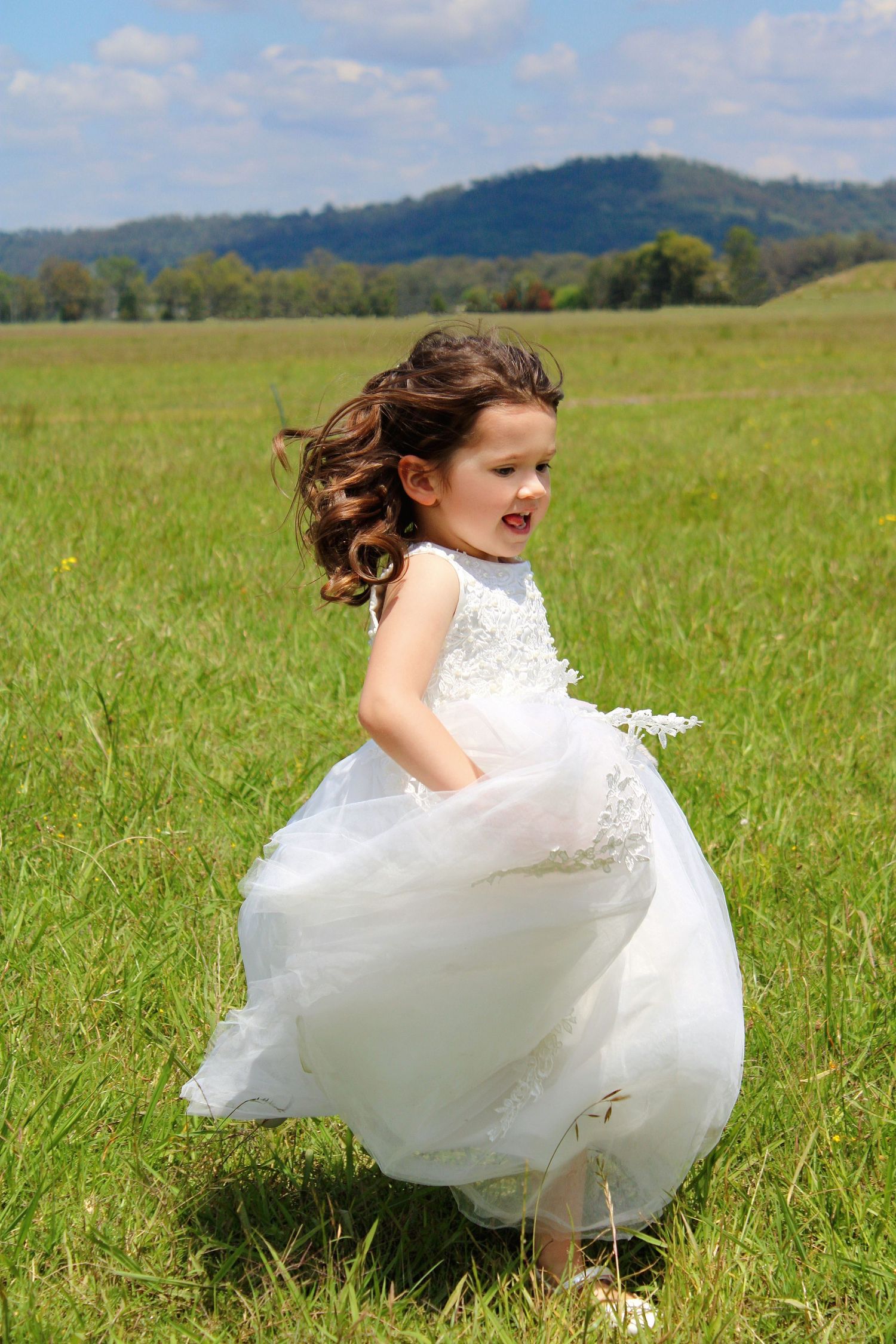 Young flower girl in white lace dress twirling in green meadow with mountains in background.