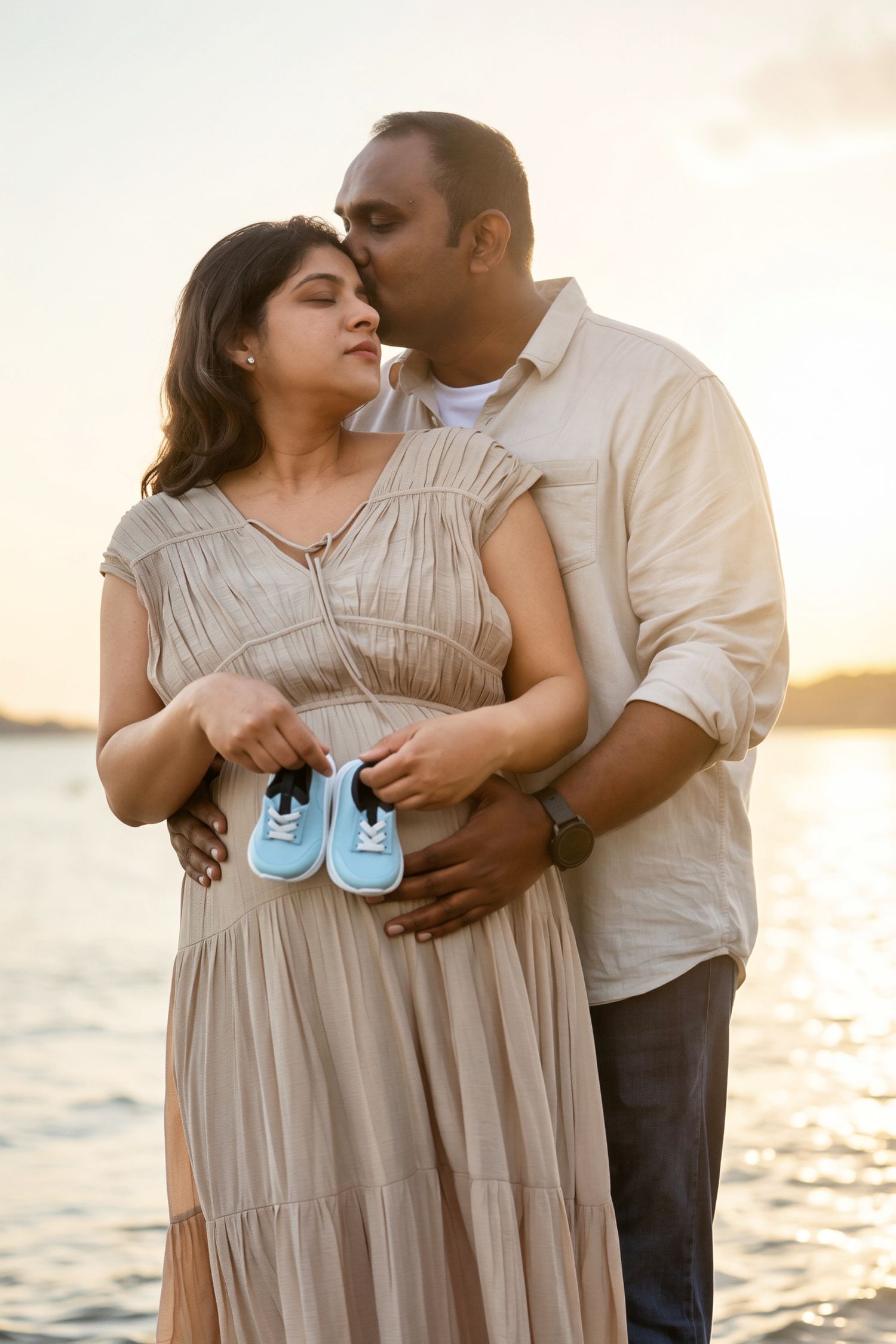Couple at sunset by water, man kissing pregnant woman holding tiny blue baby shoes tenderly.