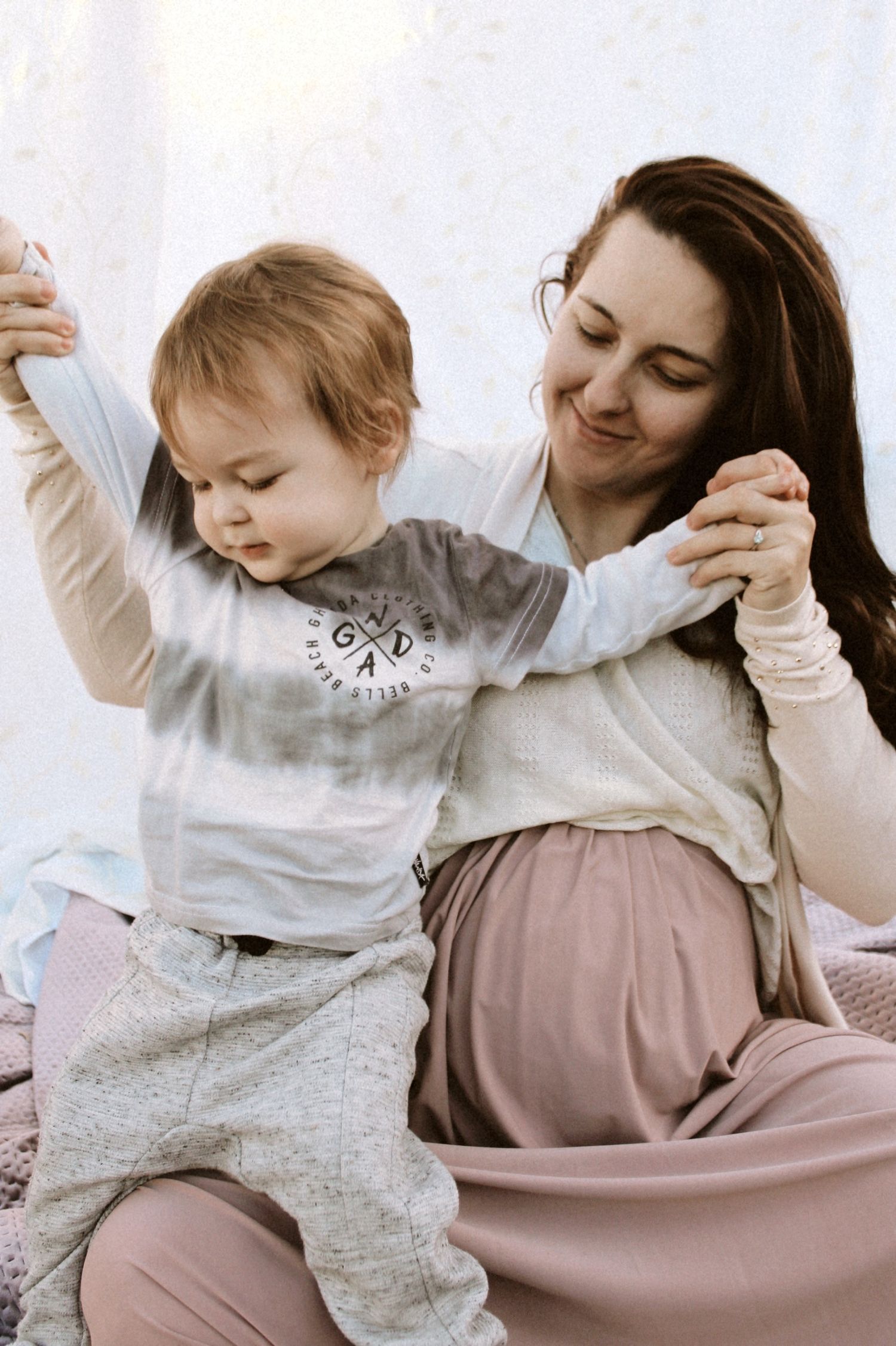 Pregnant mother smiling while helping her toddler raise arms during an indoor maternity photoshoot.
