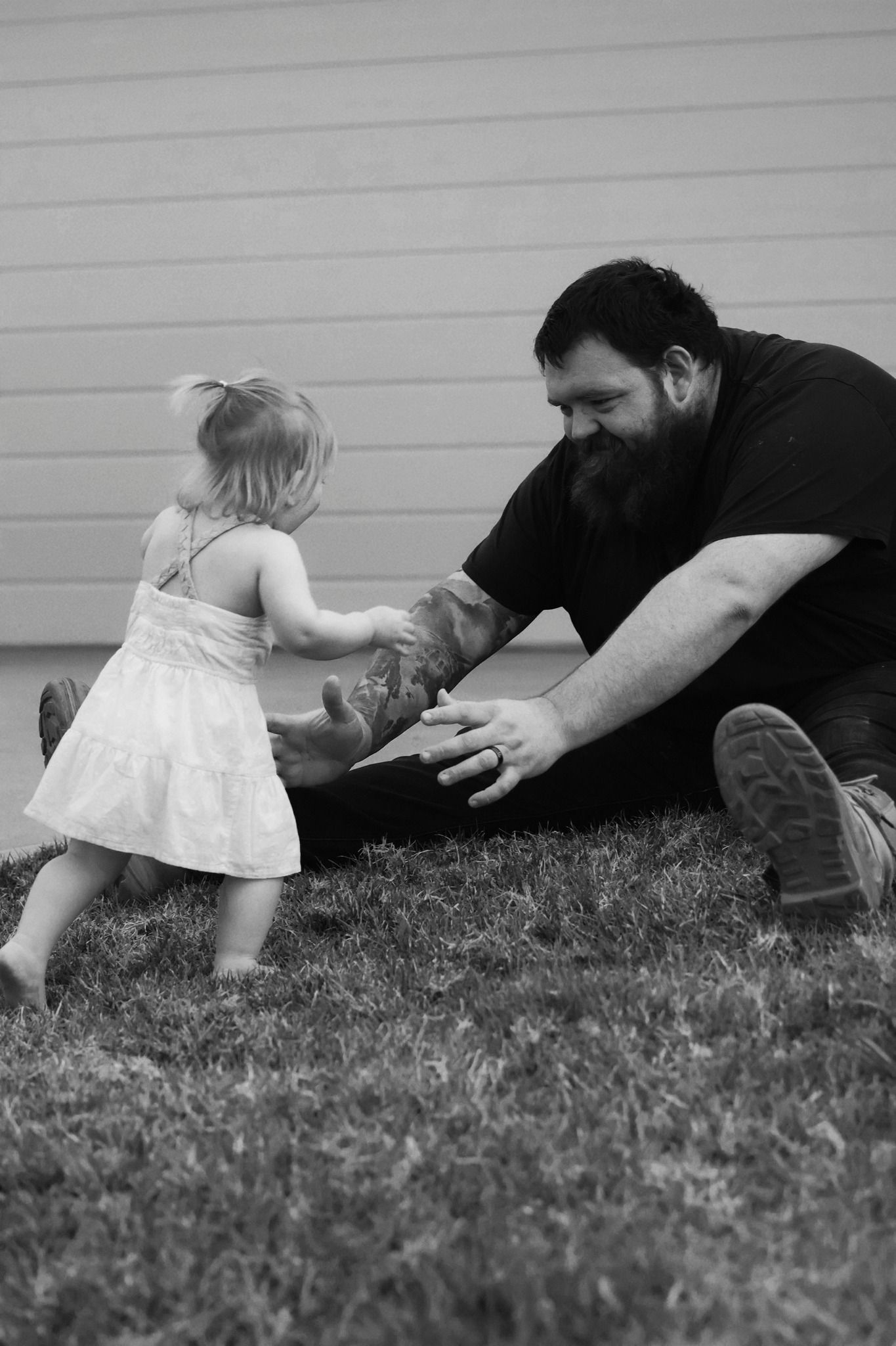 Bearded dad kneeling on grass playing with young daughter in white dress, black and white photo.