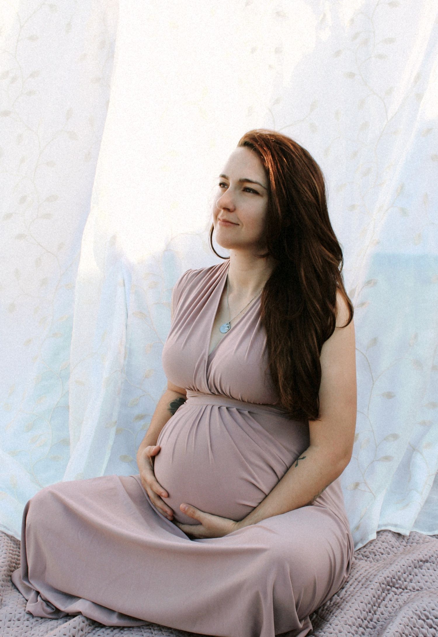 Beautiful pregnant woman in mauve dress sitting gracefully holding baby bump near flowing white curtains.
