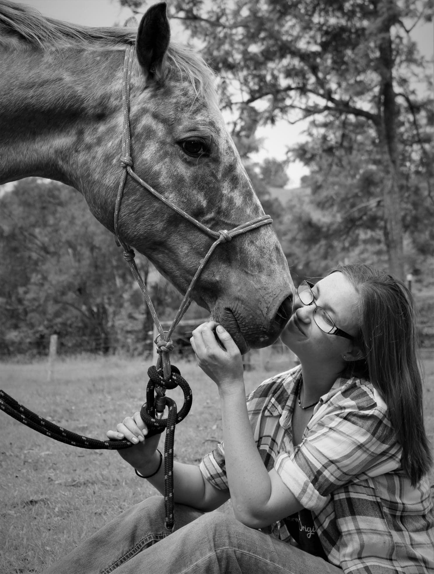 Black and white photo of a young woman in glasses sharing a tender moment kissing a horse outdoors.