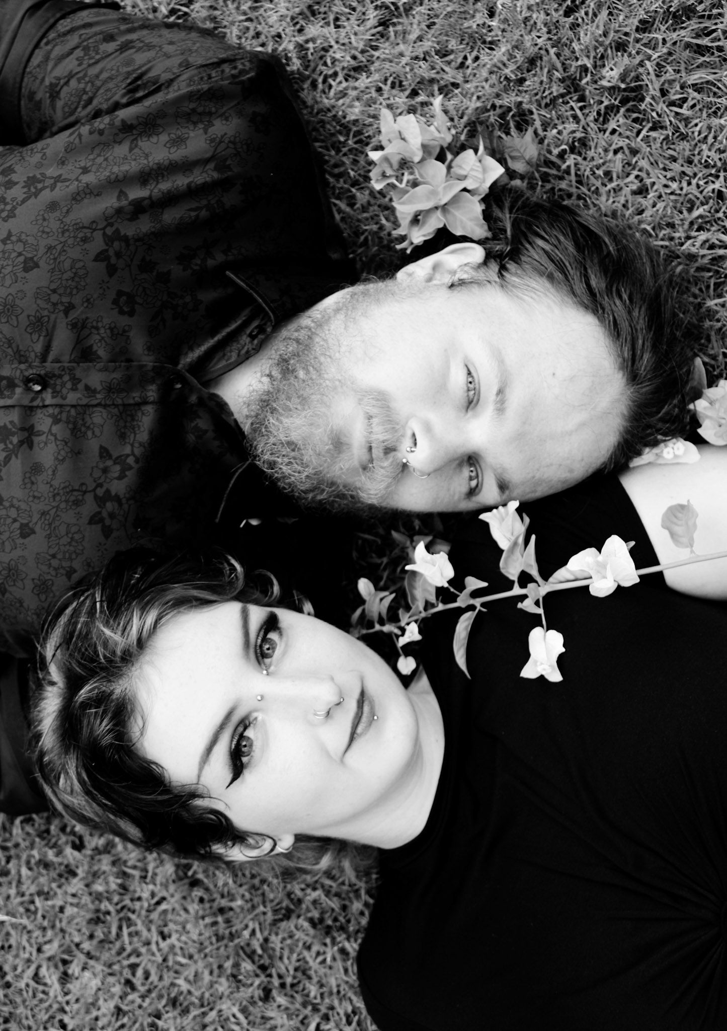 Black and white overhead portrait of couple lying on grass with flowers between them.