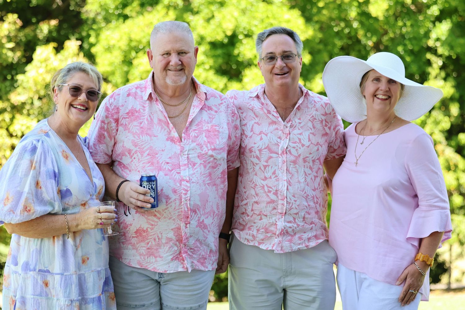 Four smiling adults posing together outdoors wearing pink and floral summer outfits at a garden event.