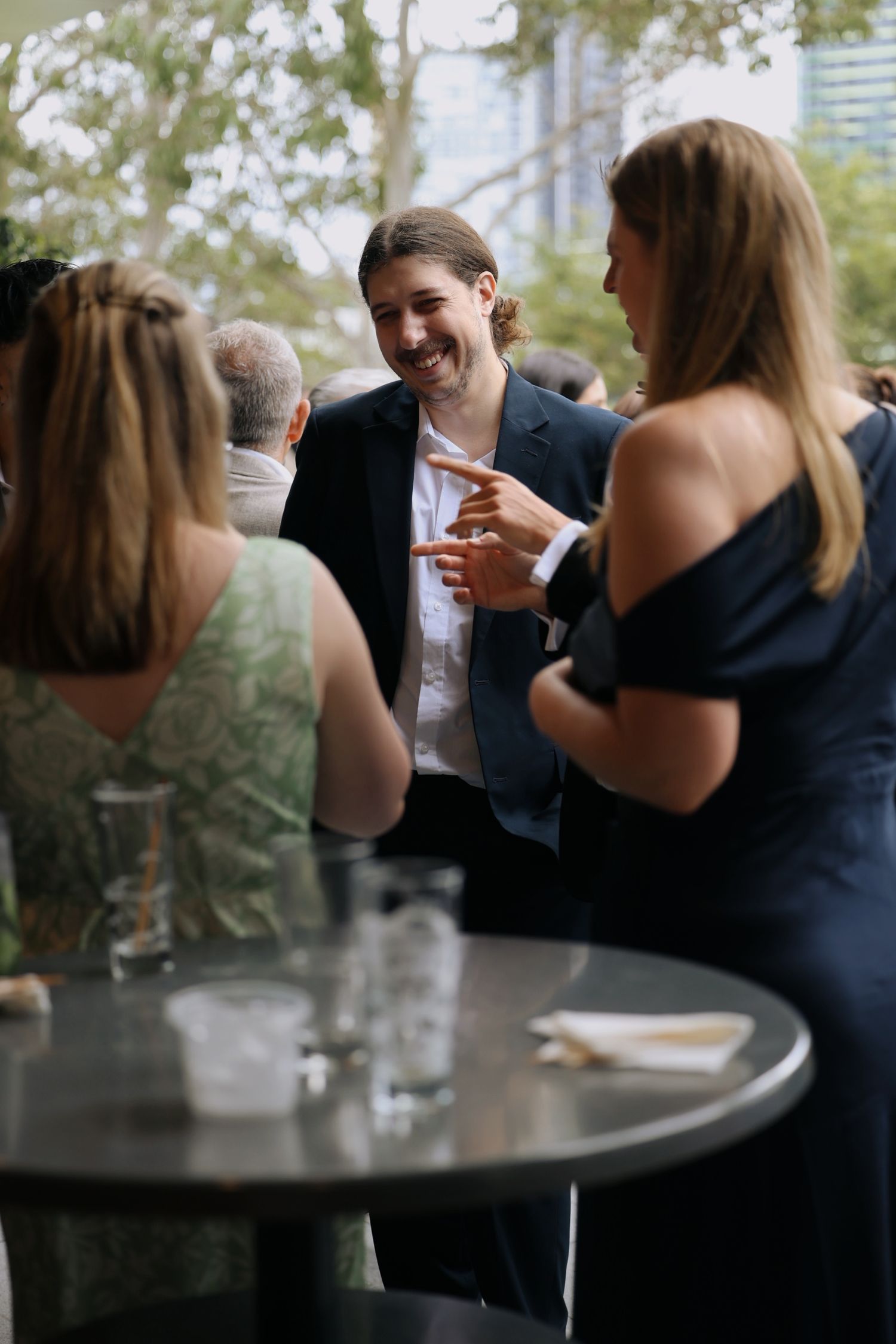 Young man in navy blazer laughing with guests at an outdoor cocktail reception.