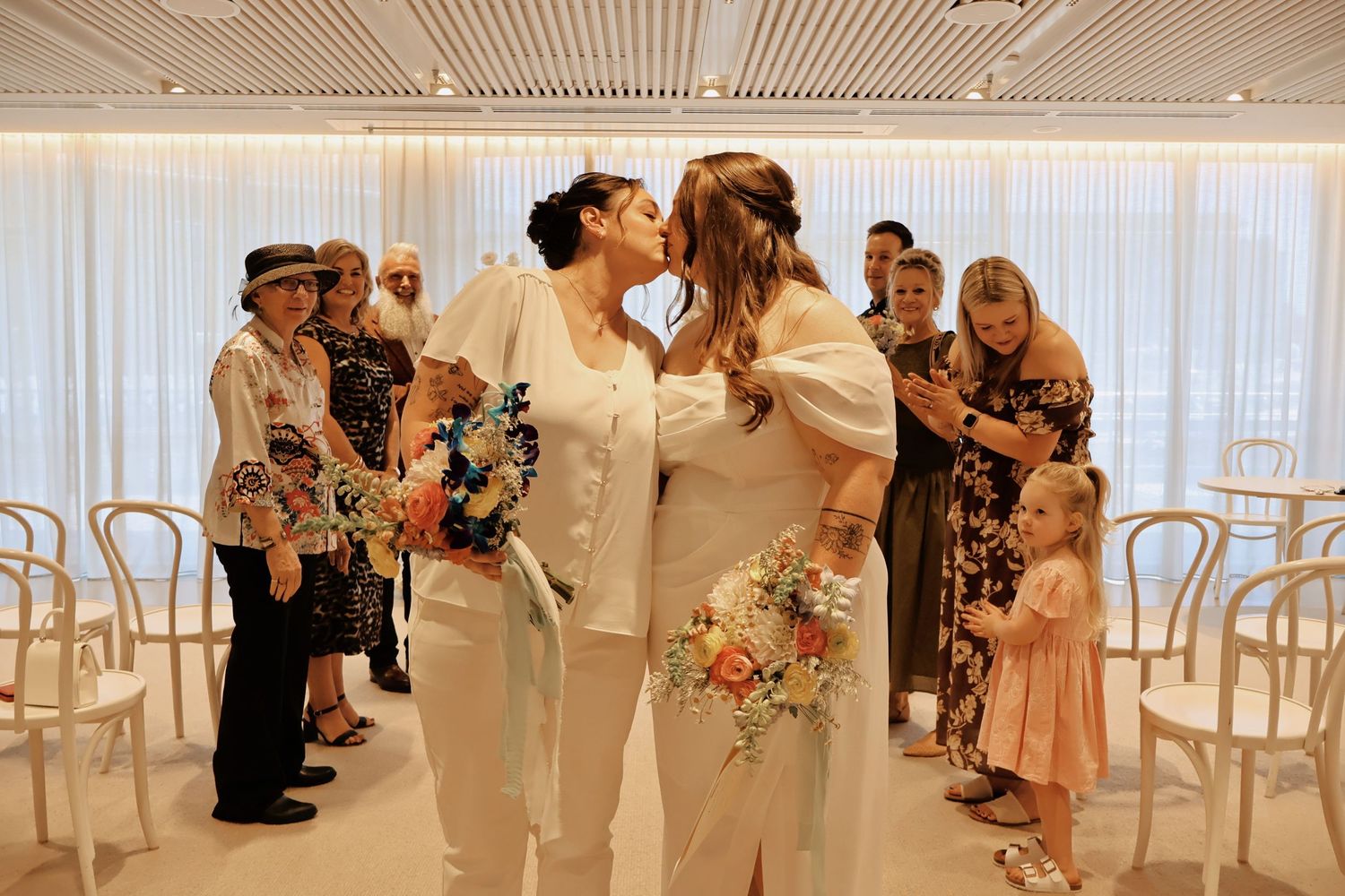 Two brides share their first kiss as a married couple surrounded by guests in a bright white venue.