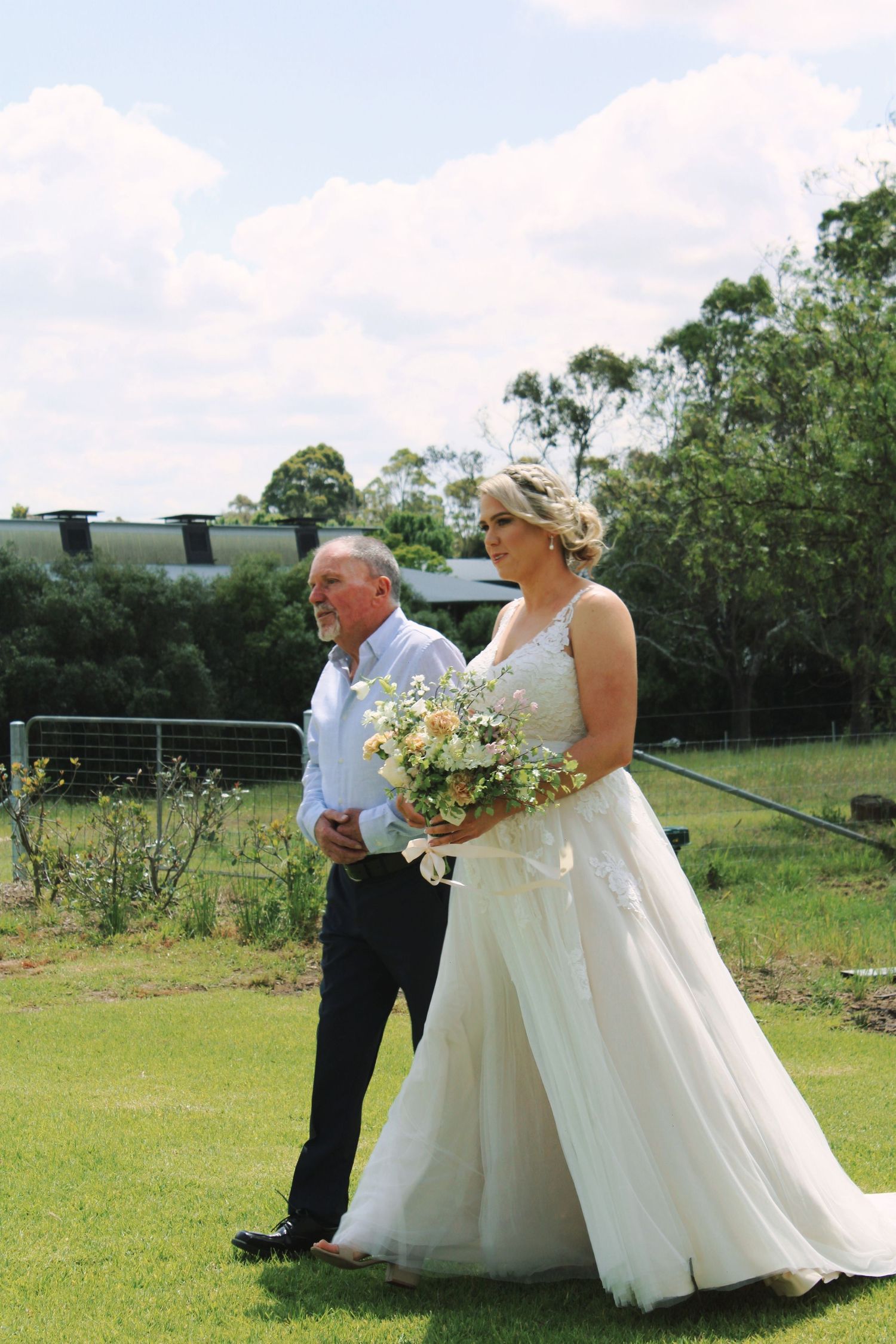 Bride in lace wedding gown holding a wildflower bouquet walks with her father across a green lawn.