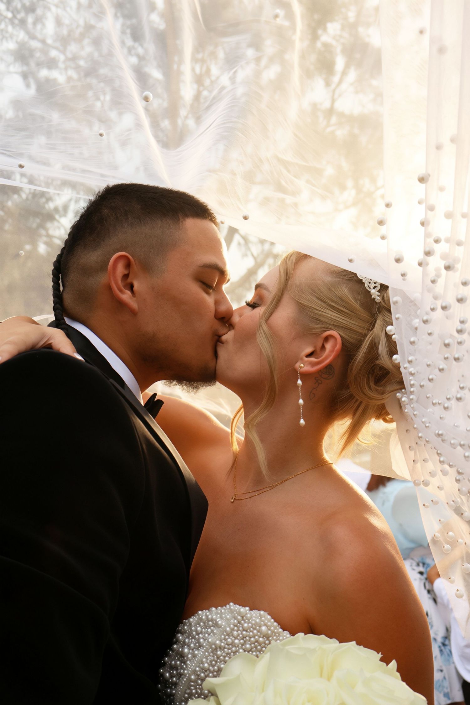 Bride and groom share a romantic kiss under a pearl-adorned veil in soft golden sunlight.