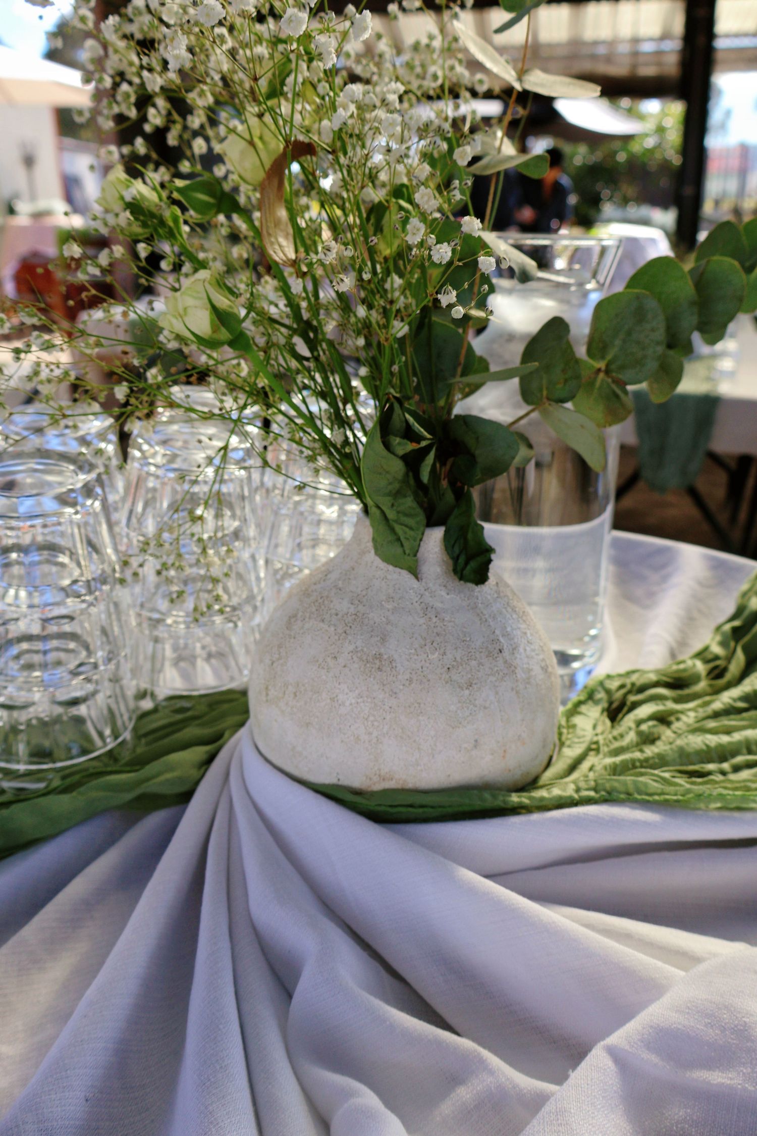 Elegant wedding table centerpiece with white stone vase holding greenery and baby's breath on draped white linen.