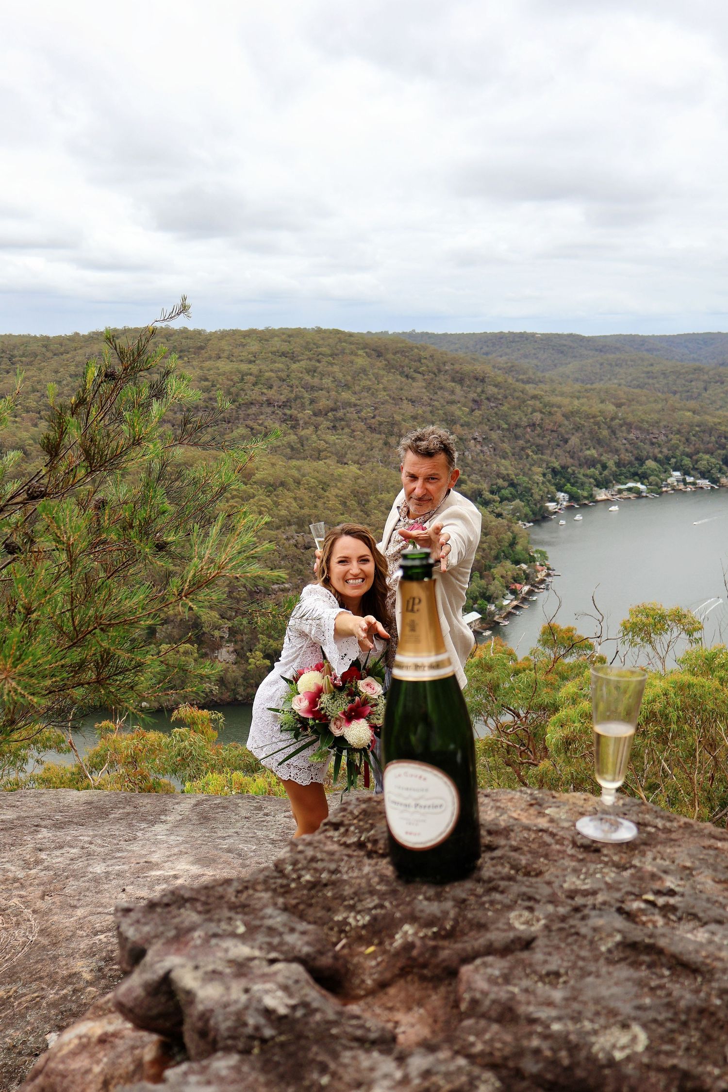 Joyful couple celebrating with champagne on a scenic cliff overlooking a river valley on their wedding day.