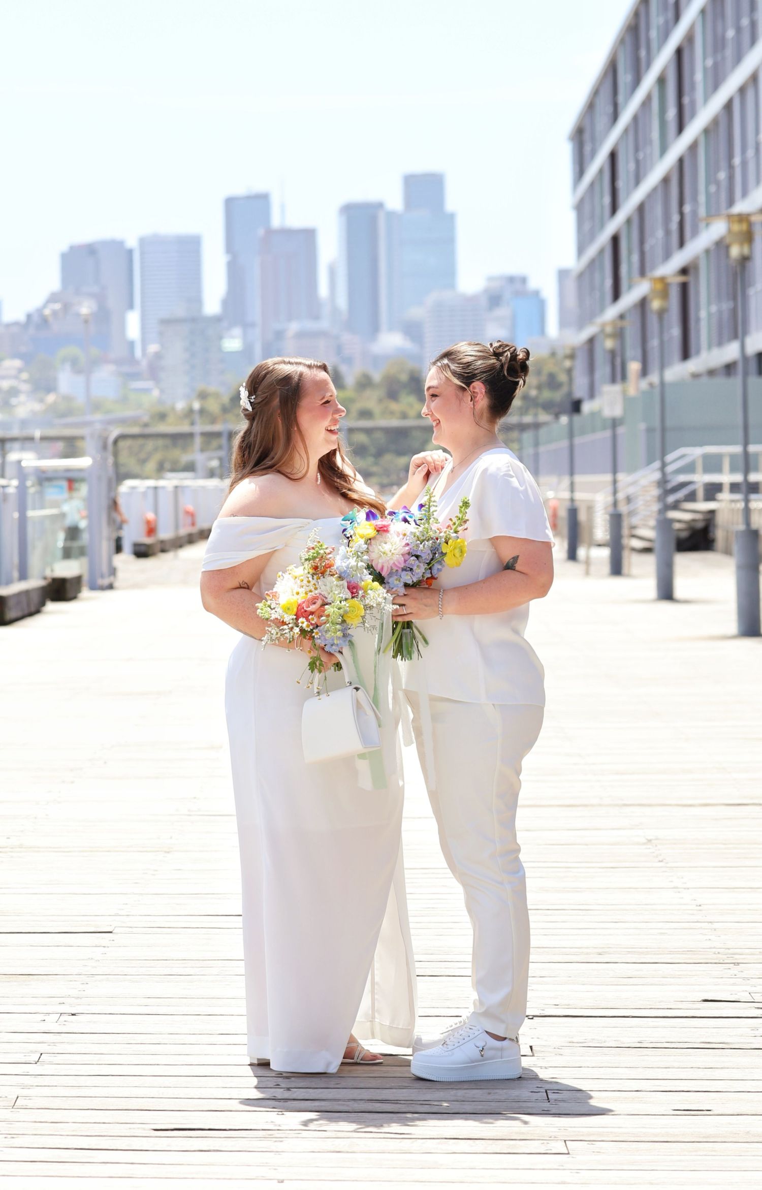 Two brides in white share a joyful moment on a sunny boardwalk with a city skyline backdrop.