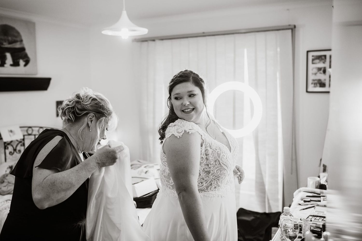 Black and white photo of a bride smiling as a woman helps her into her wedding dress in a bright room.