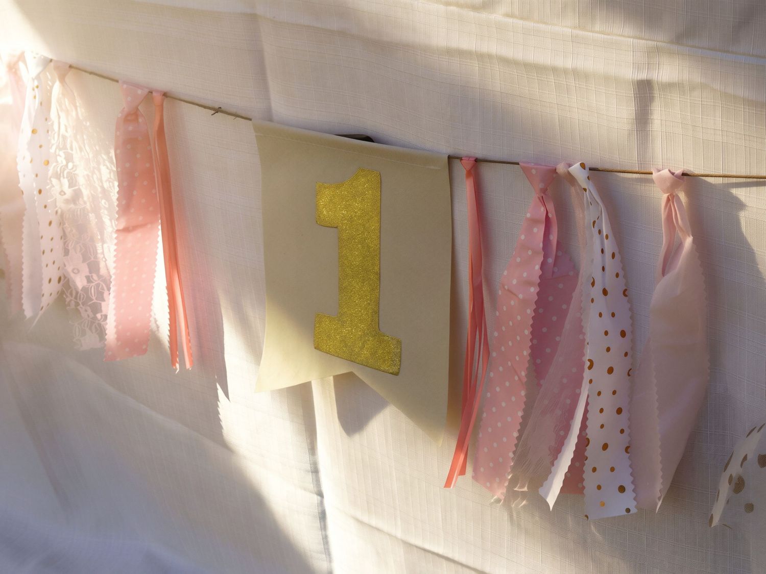 Pink and gold first birthday banner with fabric ribbons and glitter number one decoration for a party.