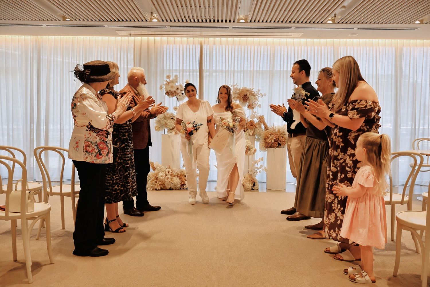 Two brides walking down aisle as guests applaud at intimate indoor wedding ceremony.