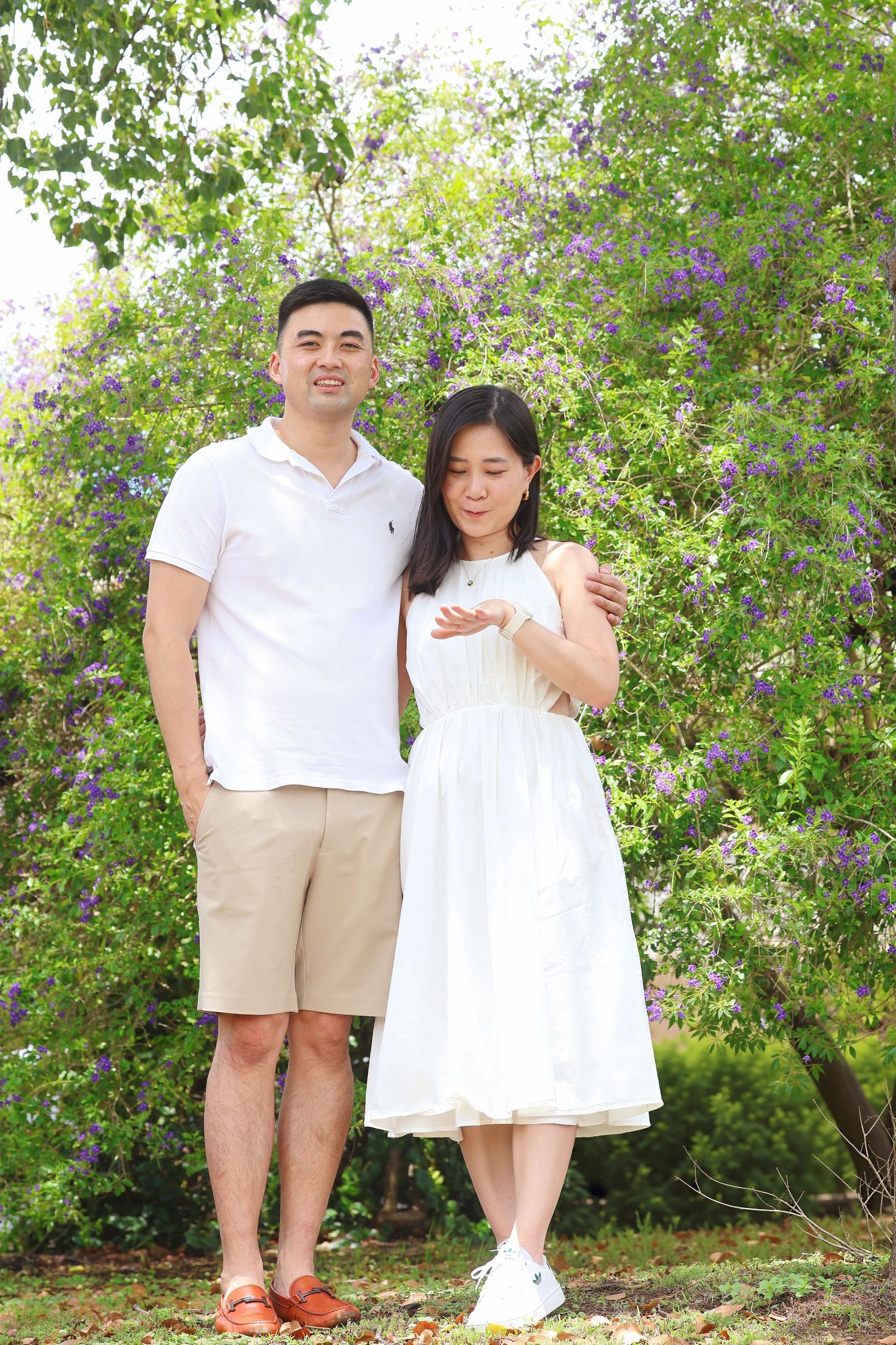 A couple stands together in a garden with purple blooming trees, both dressed casually in white and beige.