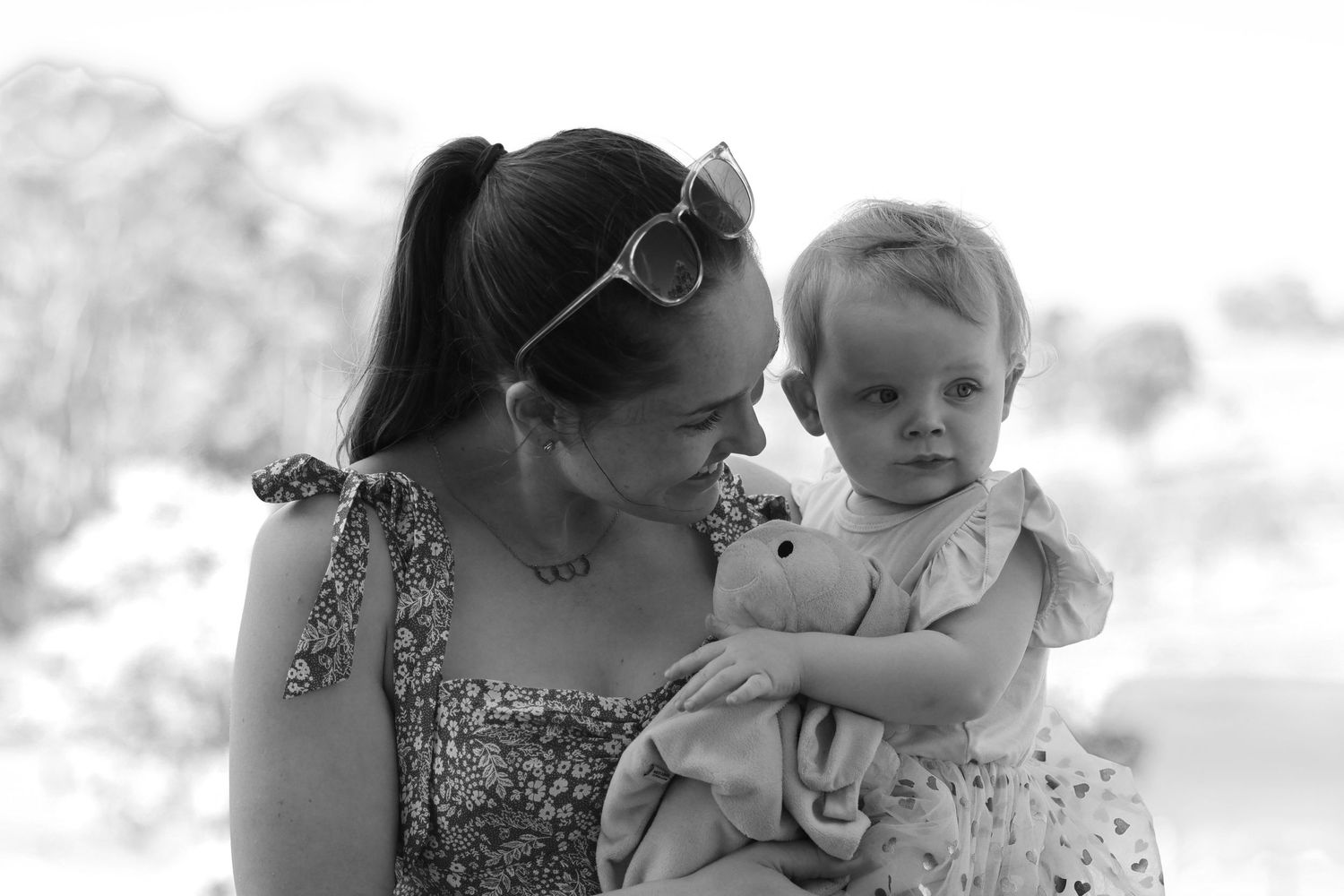 Black and white photo of a young mother holding her baby girl with a stuffed toy outdoors.