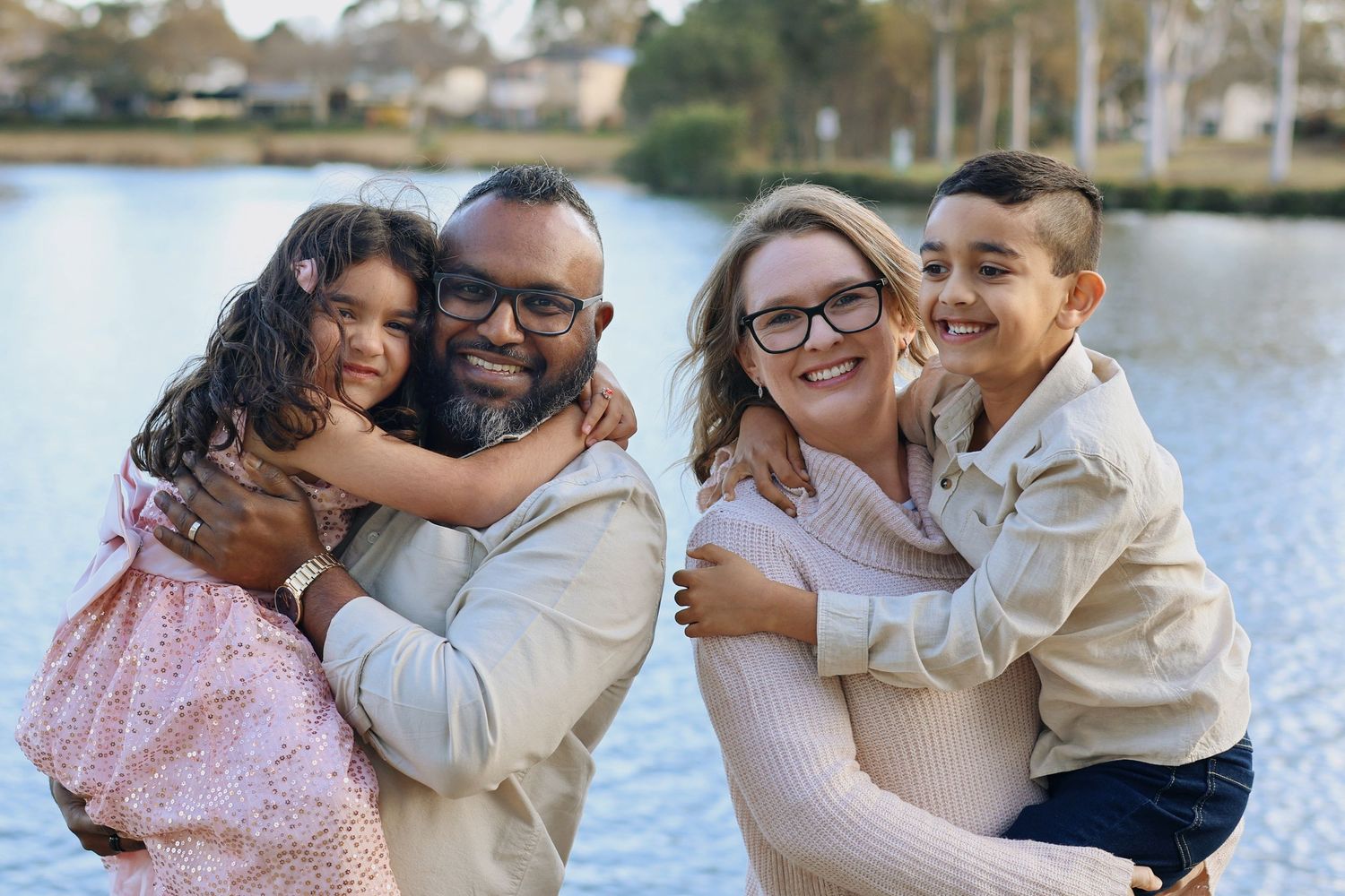 Happy family of four smiling together outdoors near a lake on a sunny day.