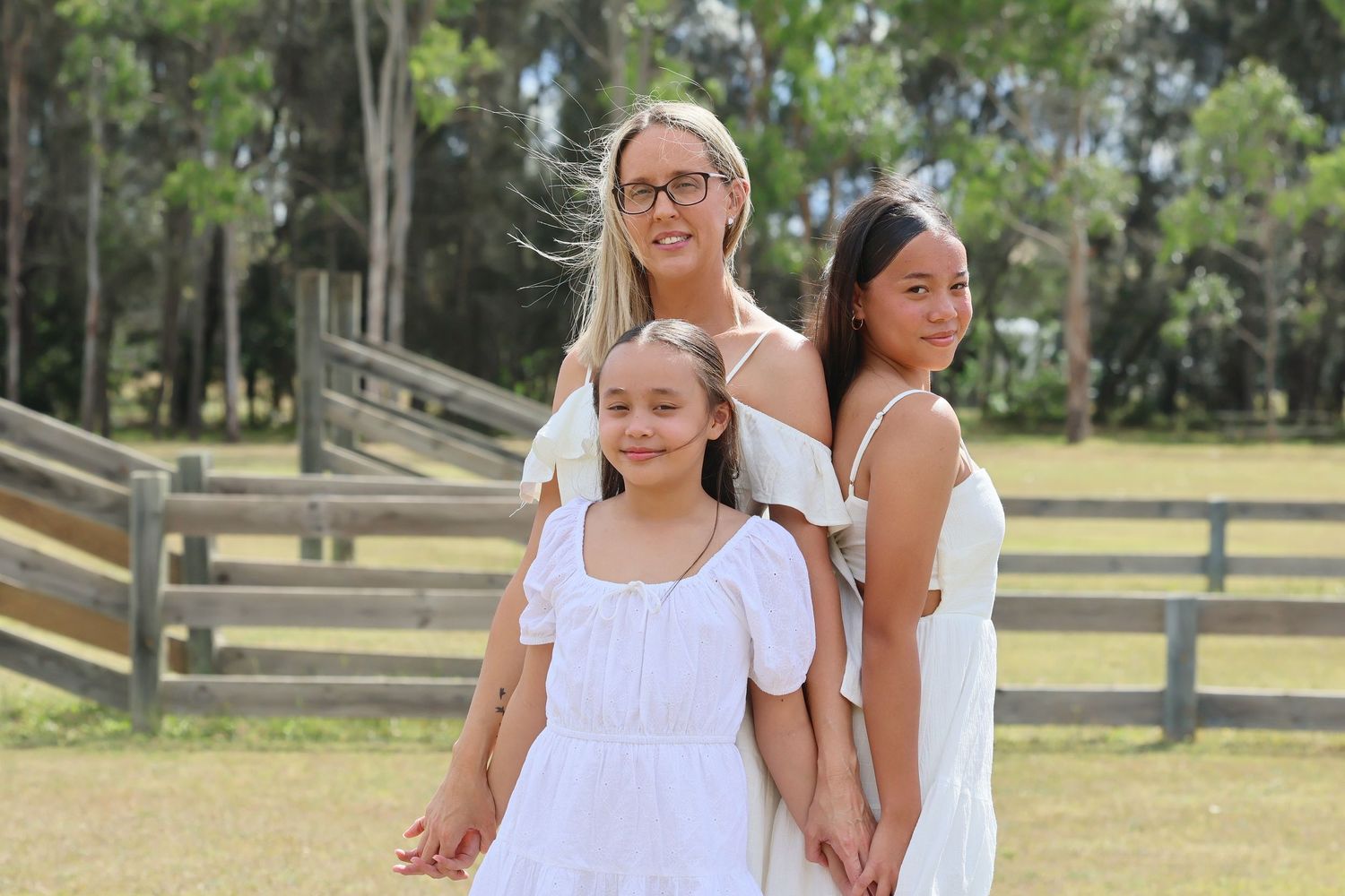 Mother and two daughters posing together in white dresses in a sunny outdoor park setting.
