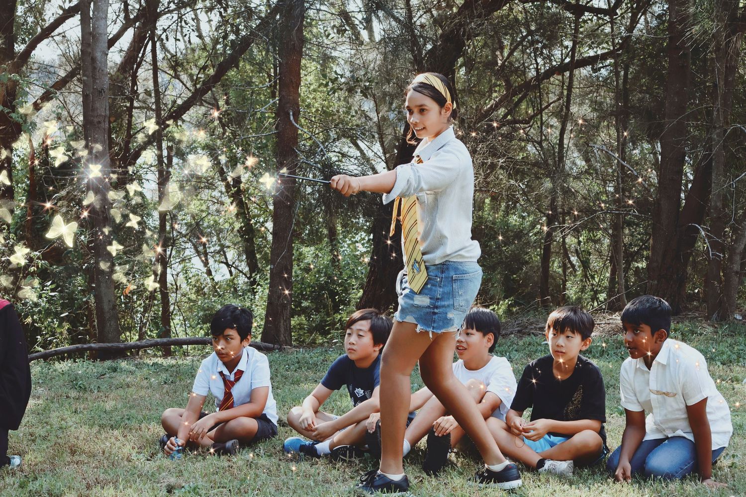 Female camp counselor leading outdoor activity with group of children sitting on grass in wooded area.
