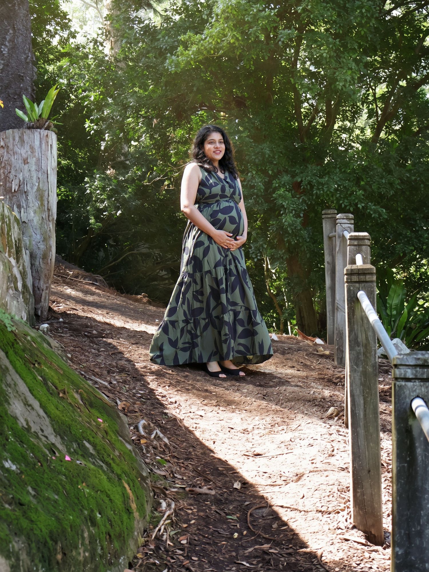 Pregnant woman in green patterned maxi dress standing on a sunlit forest path, smiling.