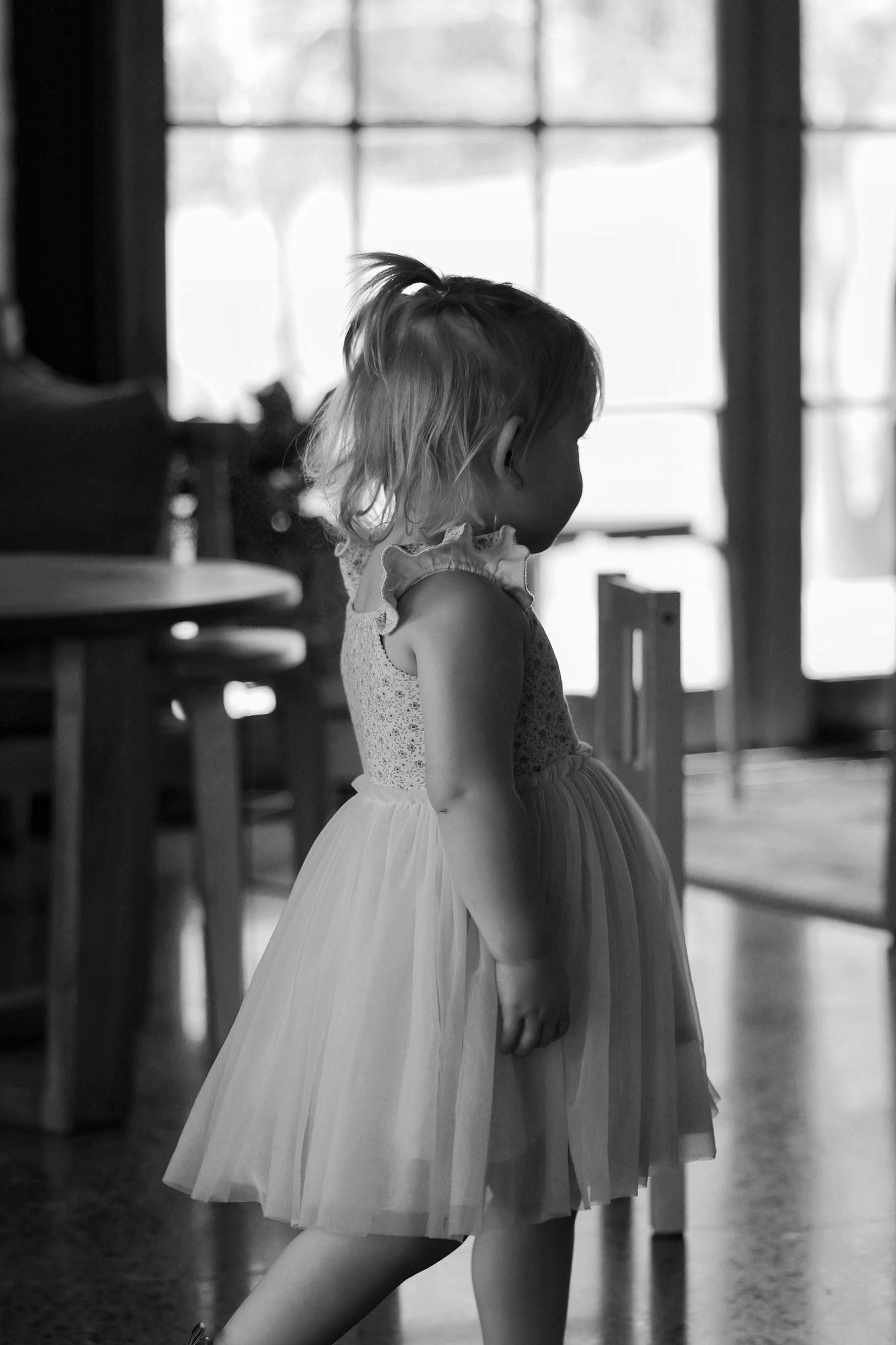 A young girl in a white tutu dress stands in a room, captured in black and white photography.