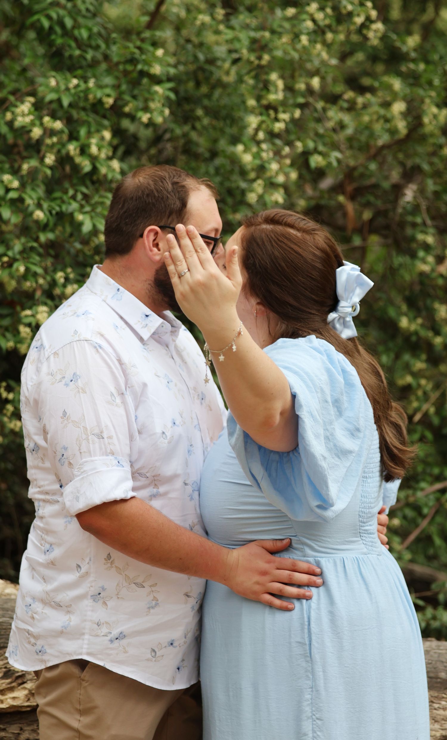 Couple shares a tender moment outdoors, bride in blue dress showing engagement ring with green trees behind.