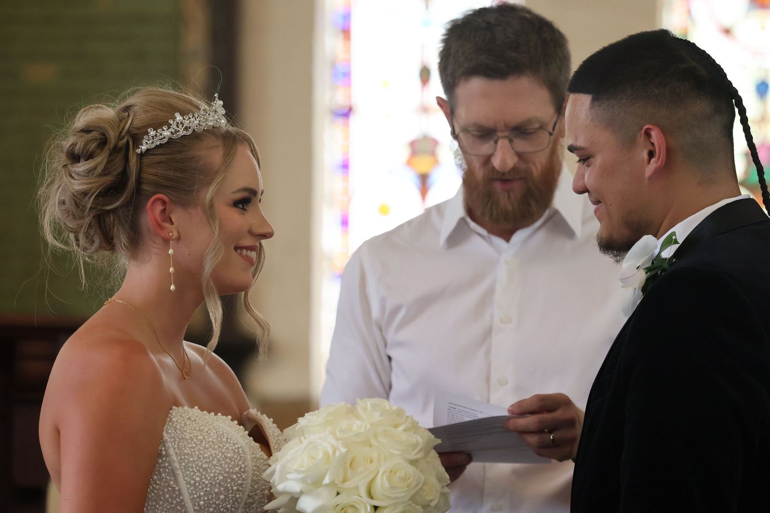 Bride smiling holding white bouquet as officiant reads vows in decorated church ceremony.