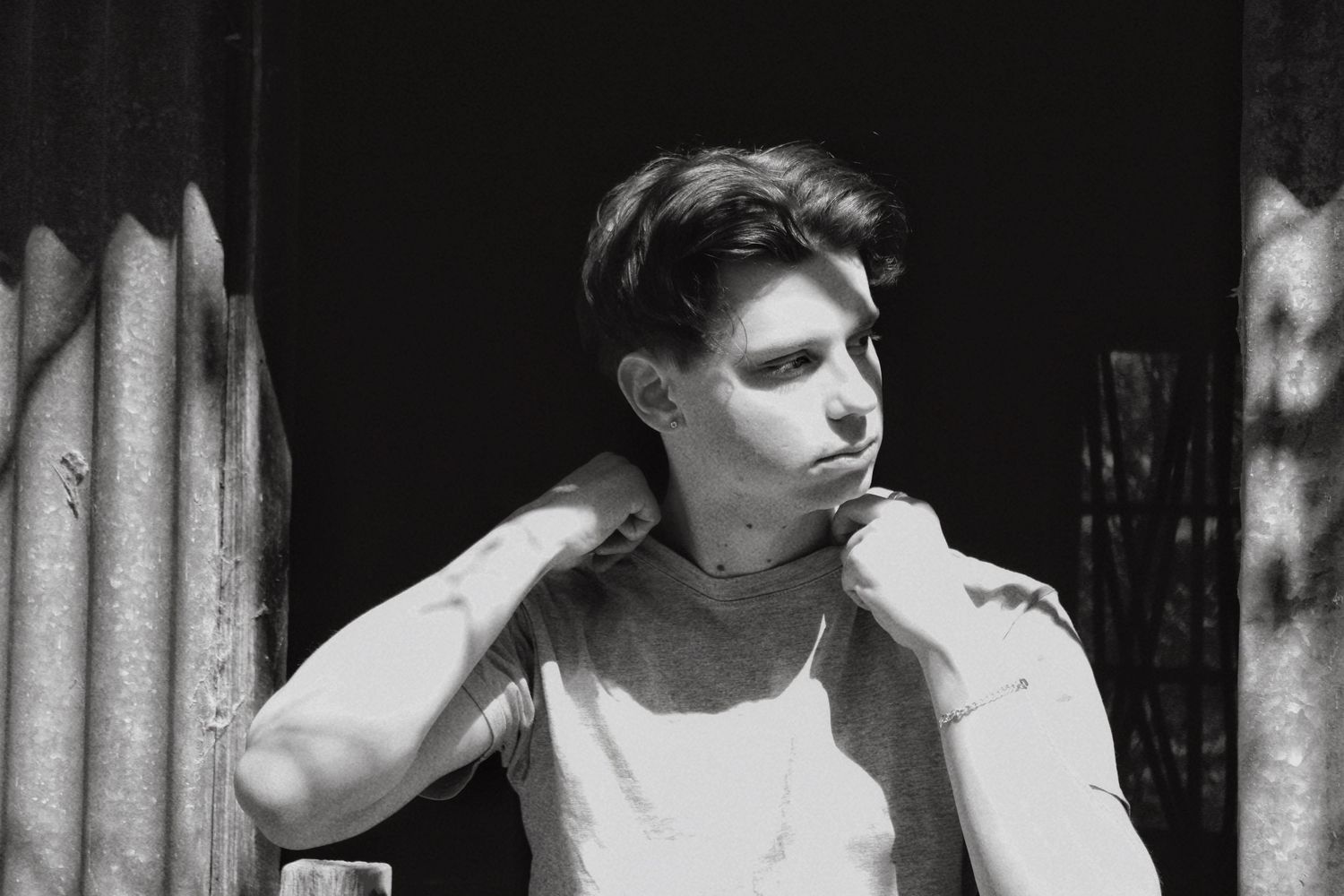 Black and white portrait of a young man in a white t-shirt leaning against a wooden structure.