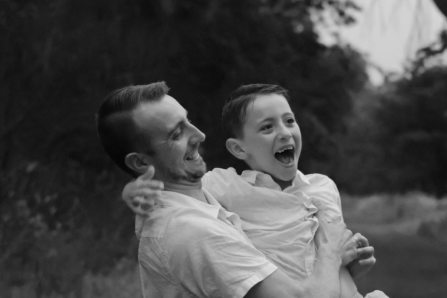 Black and white portrait of a laughing father and son sitting together in a grassy outdoor setting.
