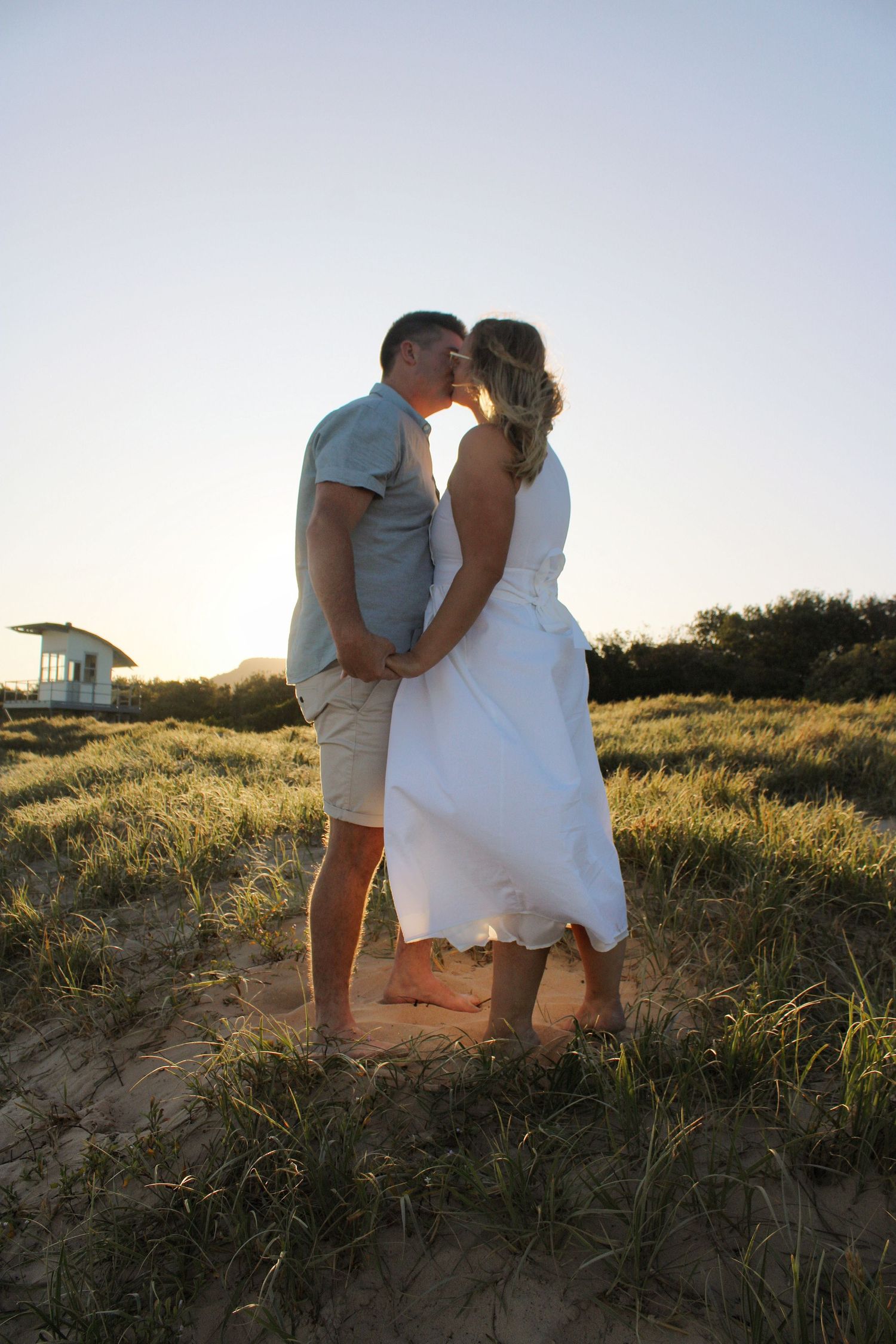 Couple kissing barefoot on sandy beach dunes at sunset, woman in white dress and man in grey shirt.