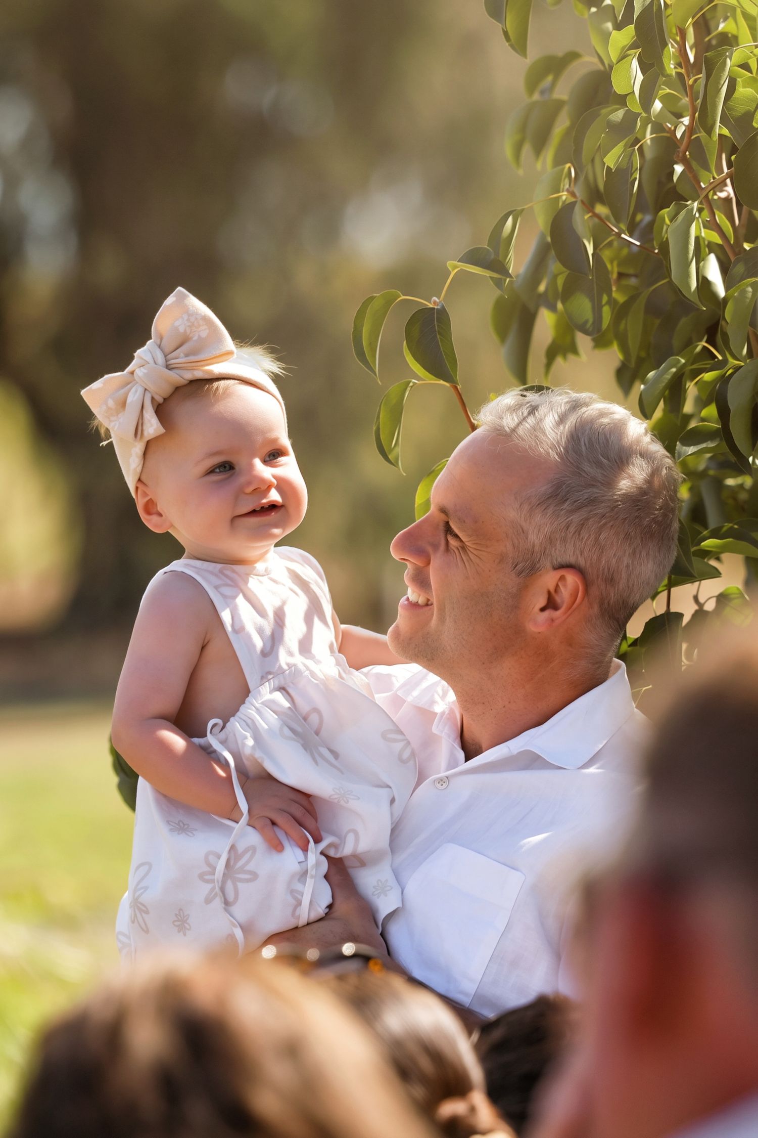 Smiling baby girl with bow headband being held by grandfather outdoors in warm golden sunlight.