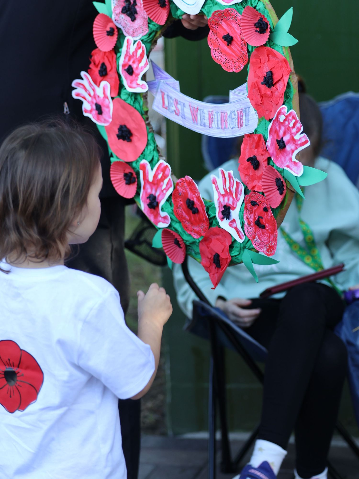Young child viewing a handmade poppy wreath with Lest We Forget banner at ANZAC ceremony.