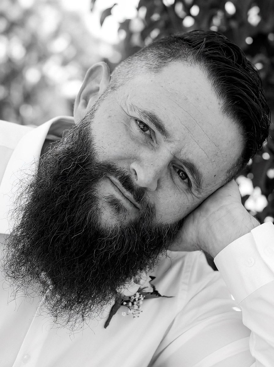 Black and white portrait of bearded groom in white shirt with boutonniere, resting head on hand.