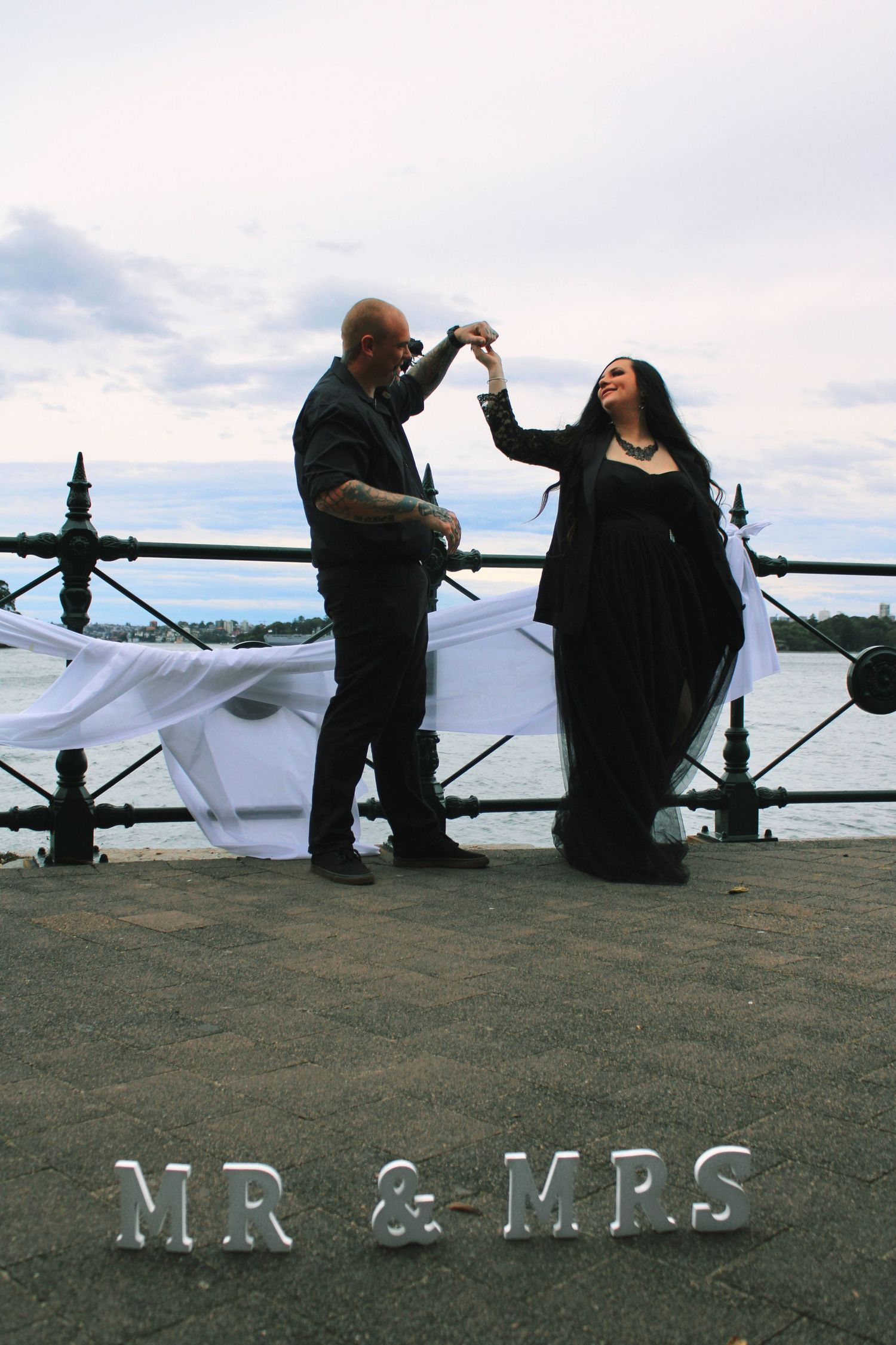 Gothic wedding couple dancing by waterfront with Mr and Mrs letter signs on ground.